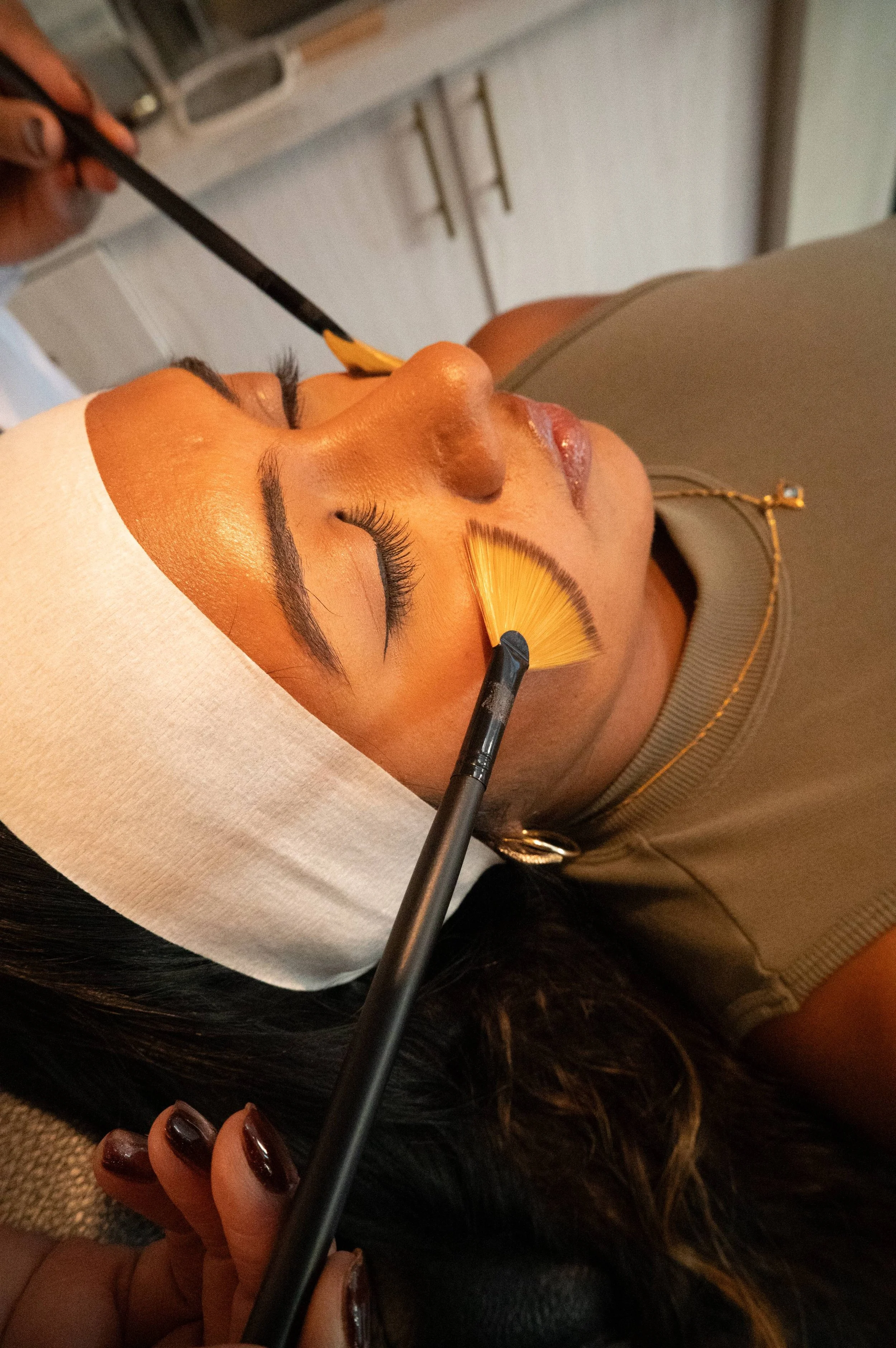 A woman lying face up receiving a facial treatment. An aesthetician using a fan brush on her face, and her eyes are closed.