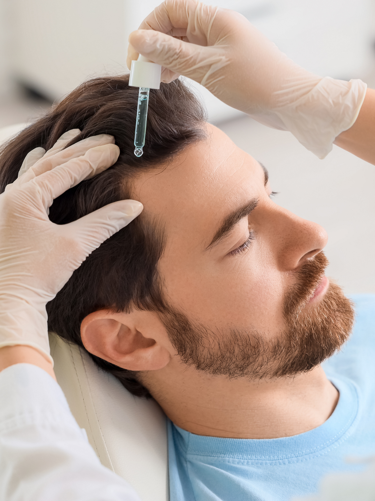 A person receiving a hair treatment with a dropper from medical gloves, lying down with eyes closed.