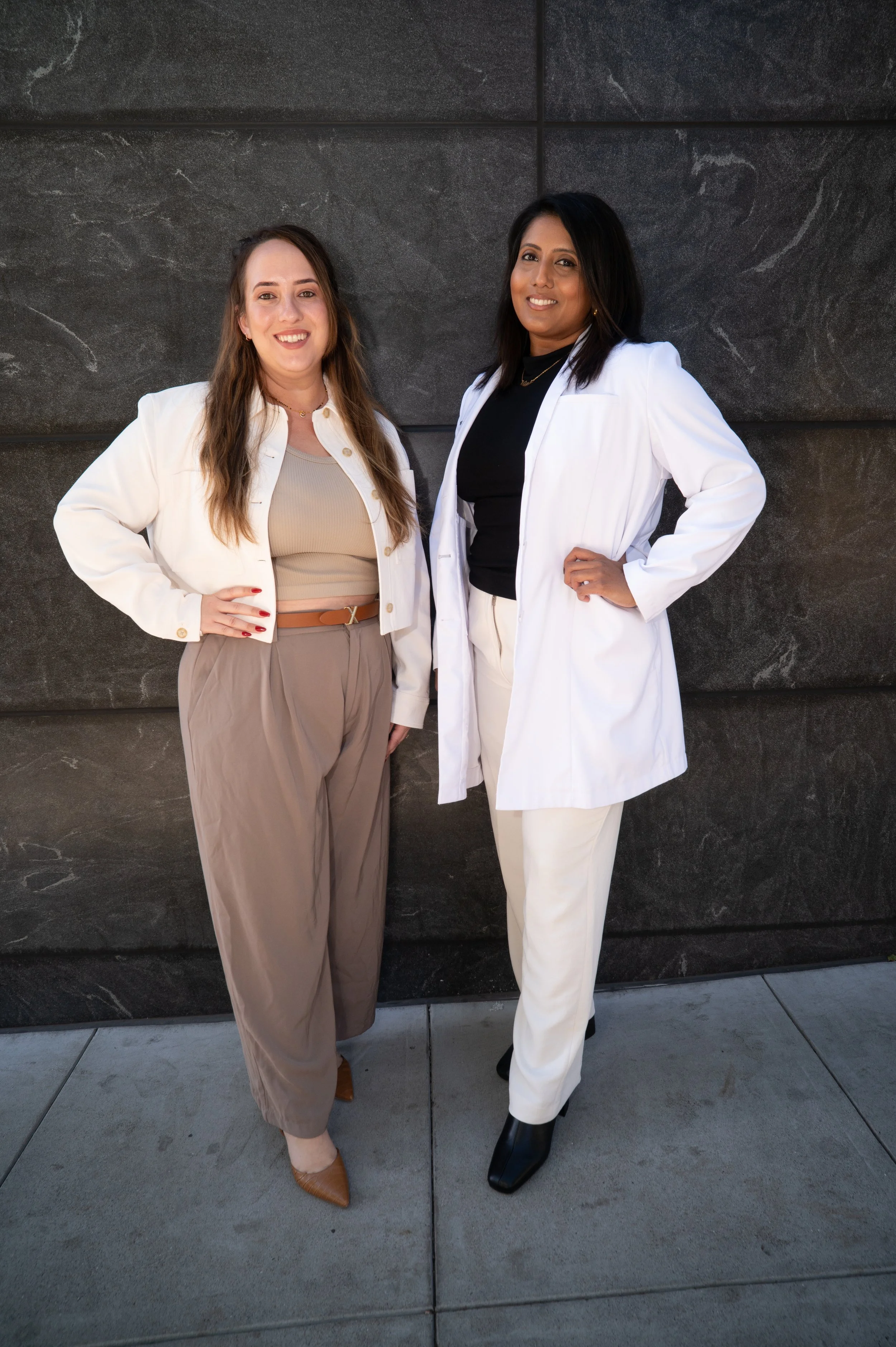 Two women standing in front of a dark gray tiled wall, smiling at the camera. The woman on the left has light skin, long brown hair, and is wearing a cream-colored jacket, beige top, and beige pants. The woman on the right has darker skin, straight black hair, and is wearing a white coat, black top, and white pants.