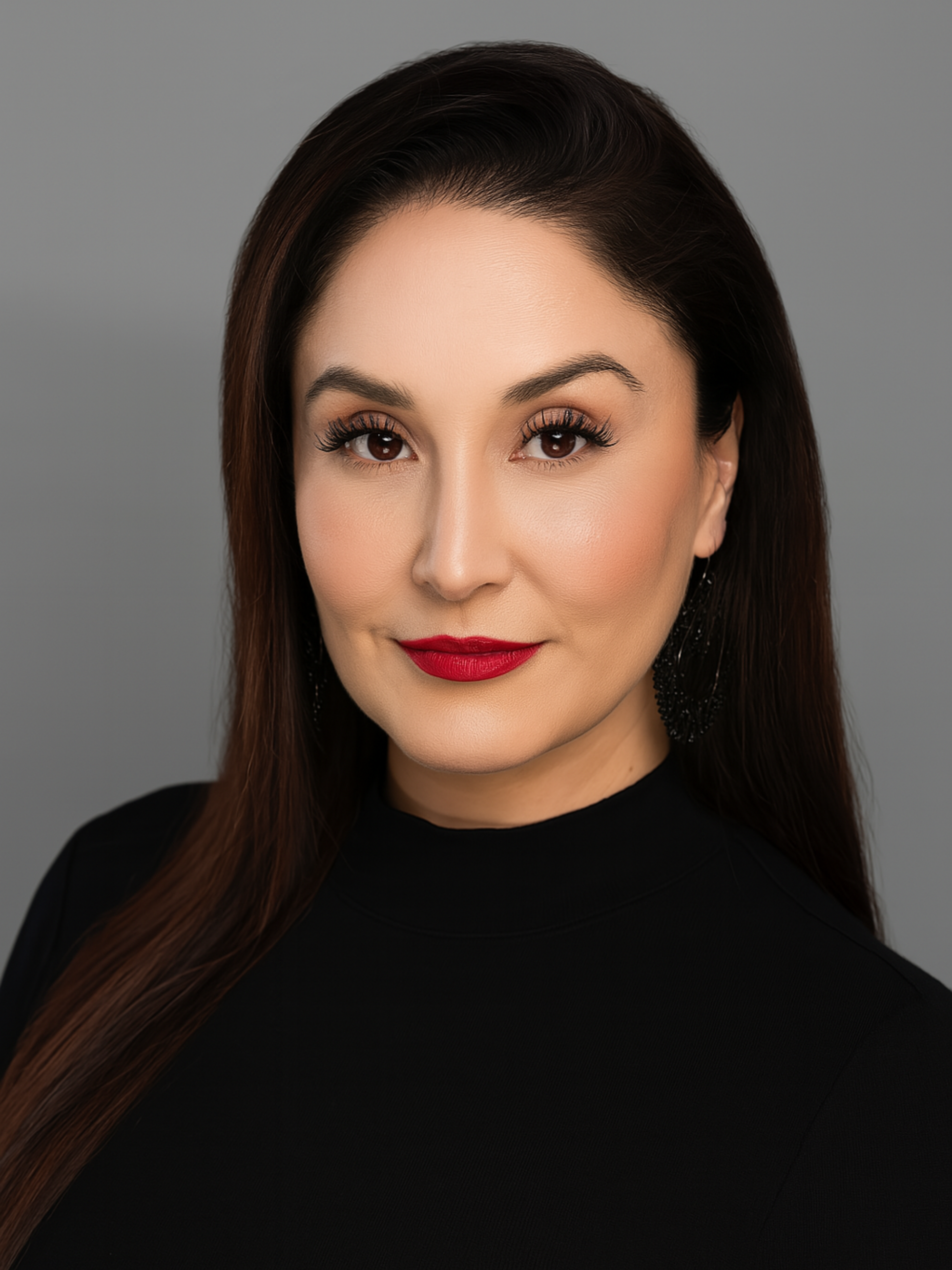 A close-up portrait of a woman with long brown hair, makeup, wearing red lipstick, black earrings, and a black top, against a gray background.