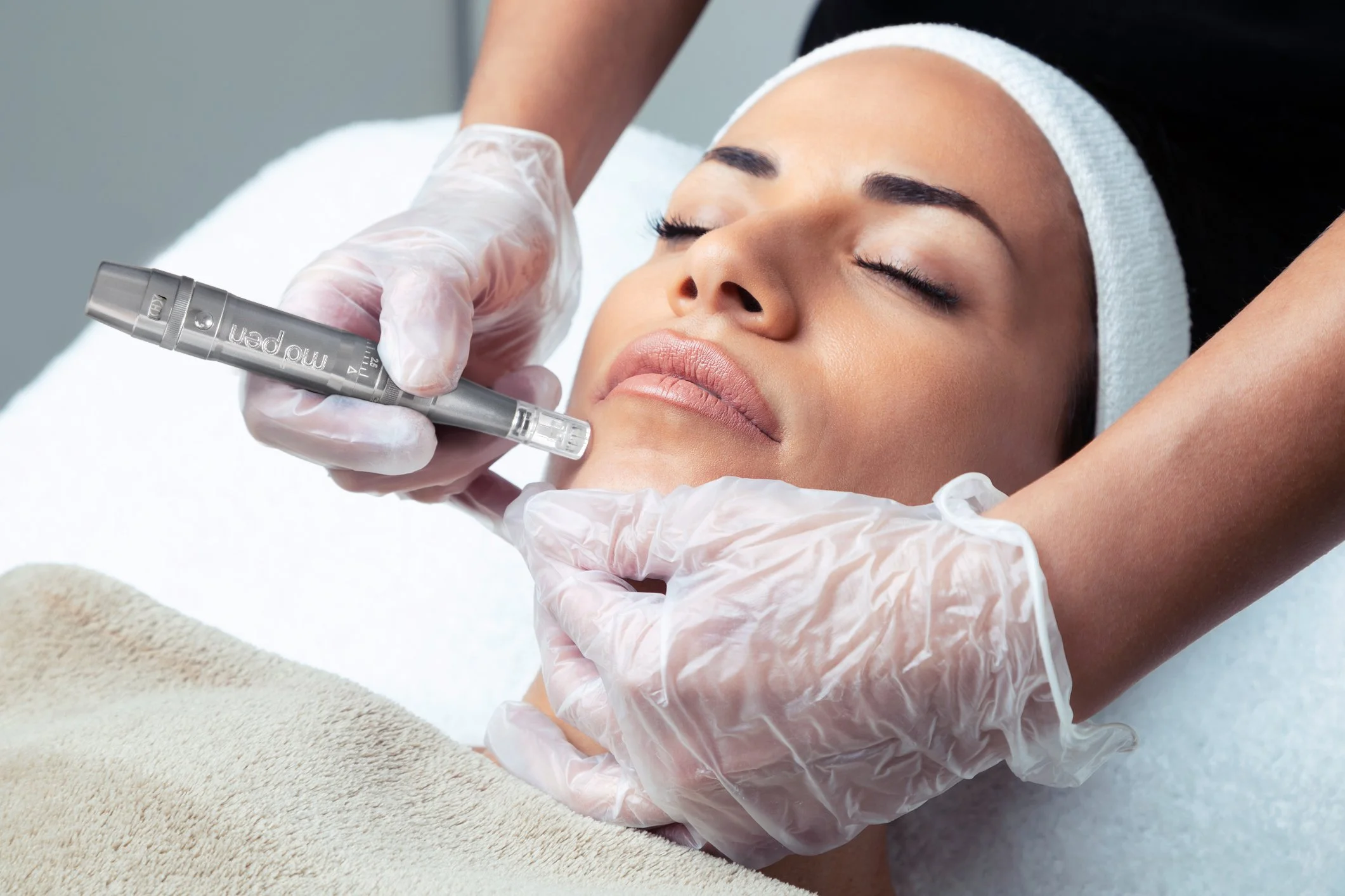 A woman receiving a facial treatment at a spa, with her eyes closed and a towel wrapped around her head, while a therapist applies a skincare product using a pen-like tool.