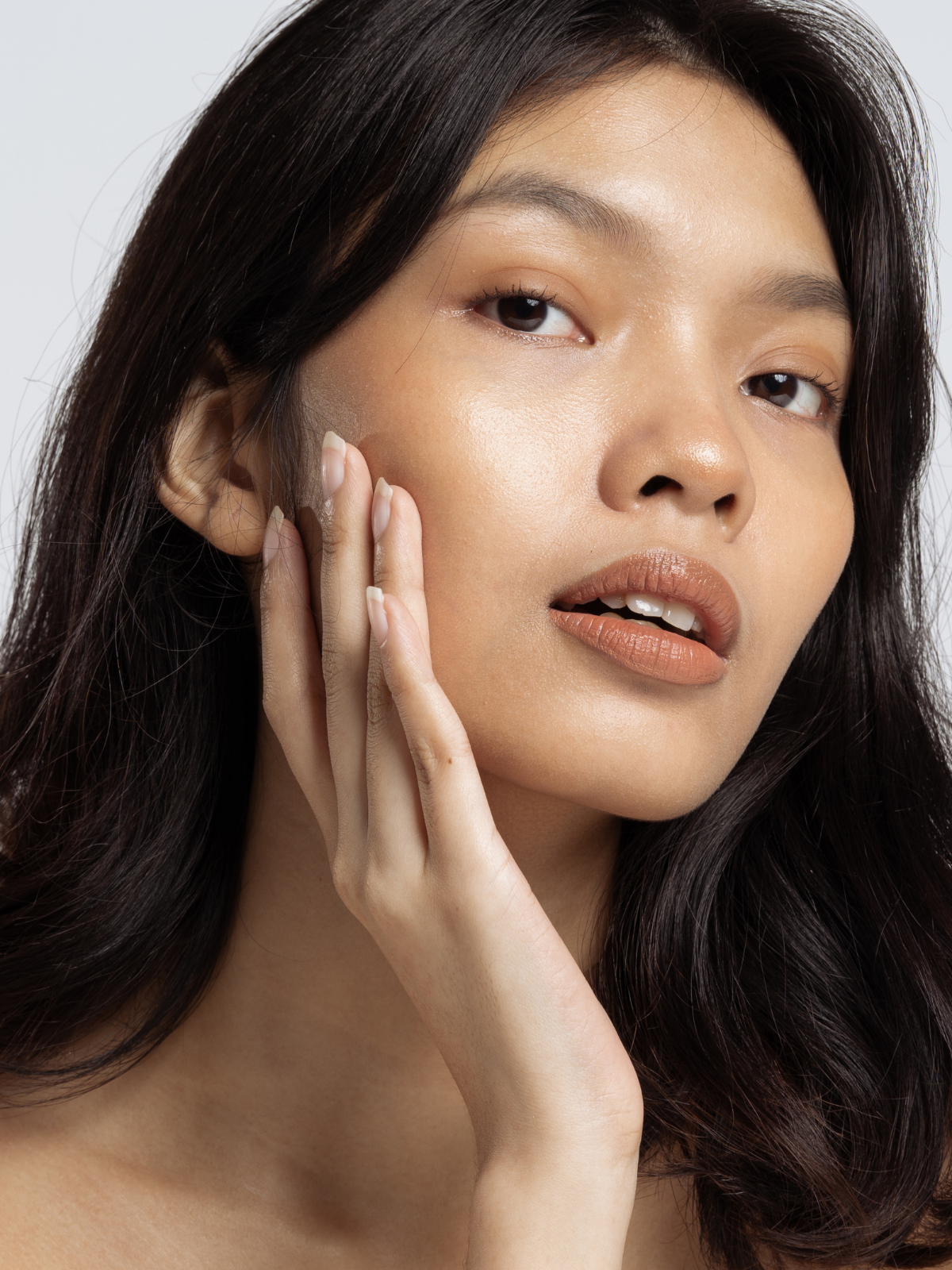 Close-up of a woman with long dark hair, lightly tanned skin, subtle makeup, and soft lips touching her face with her fingers.