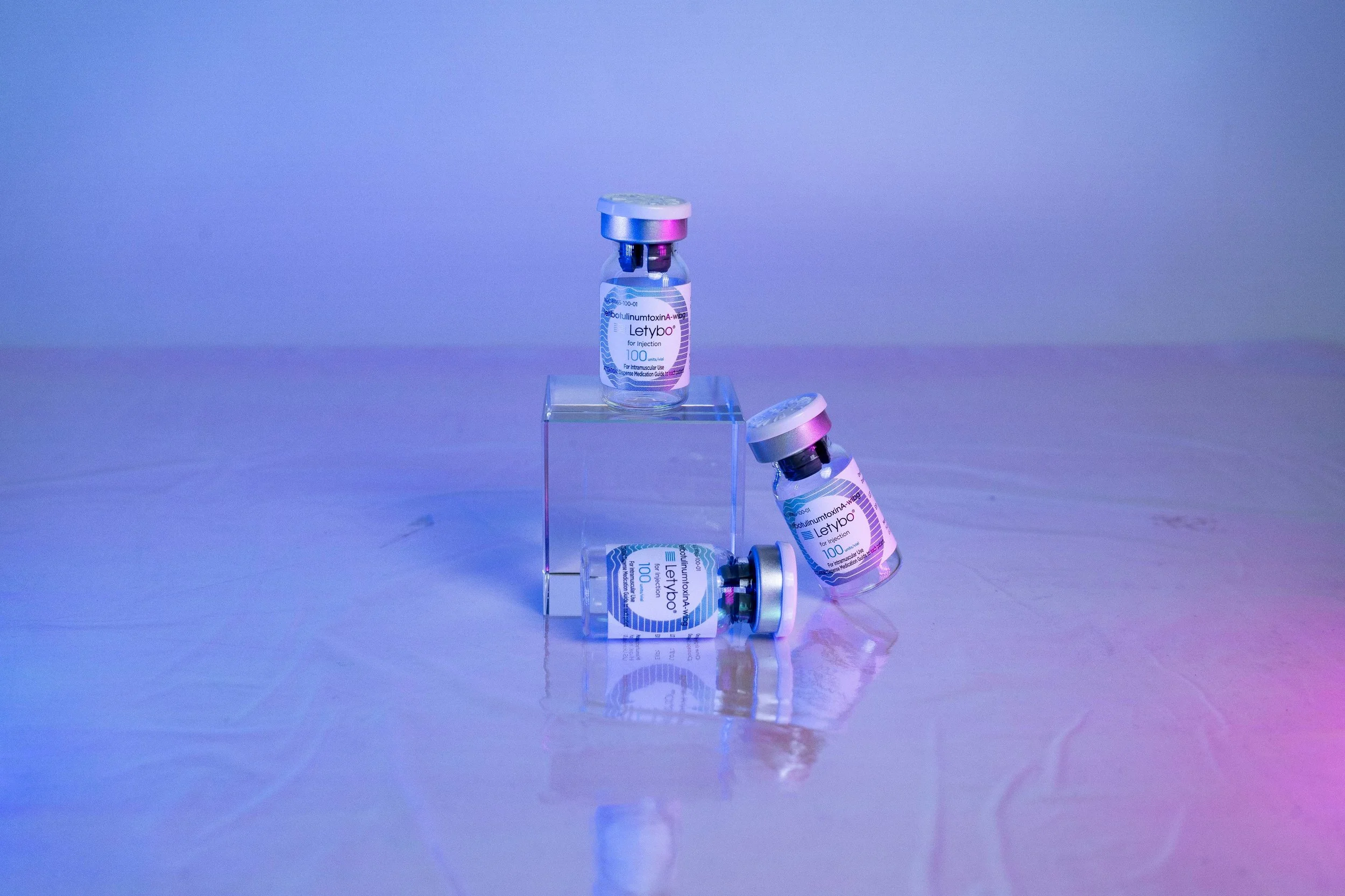 Several small vials of a medication labeled 'Letybo' are displayed. Some vials are standing upright, while others are lying down or leaning. The vials have a metallic cap and are placed on a shiny surface with a blue and purple gradient background.