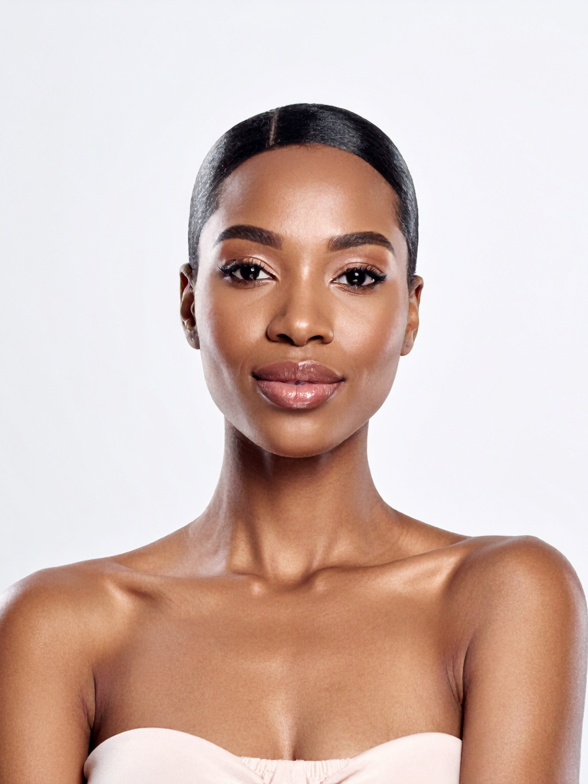 Portrait of a confident woman with smooth dark hair, minimal makeup, and nude lipstick, wearing a strapless top against a plain white background.