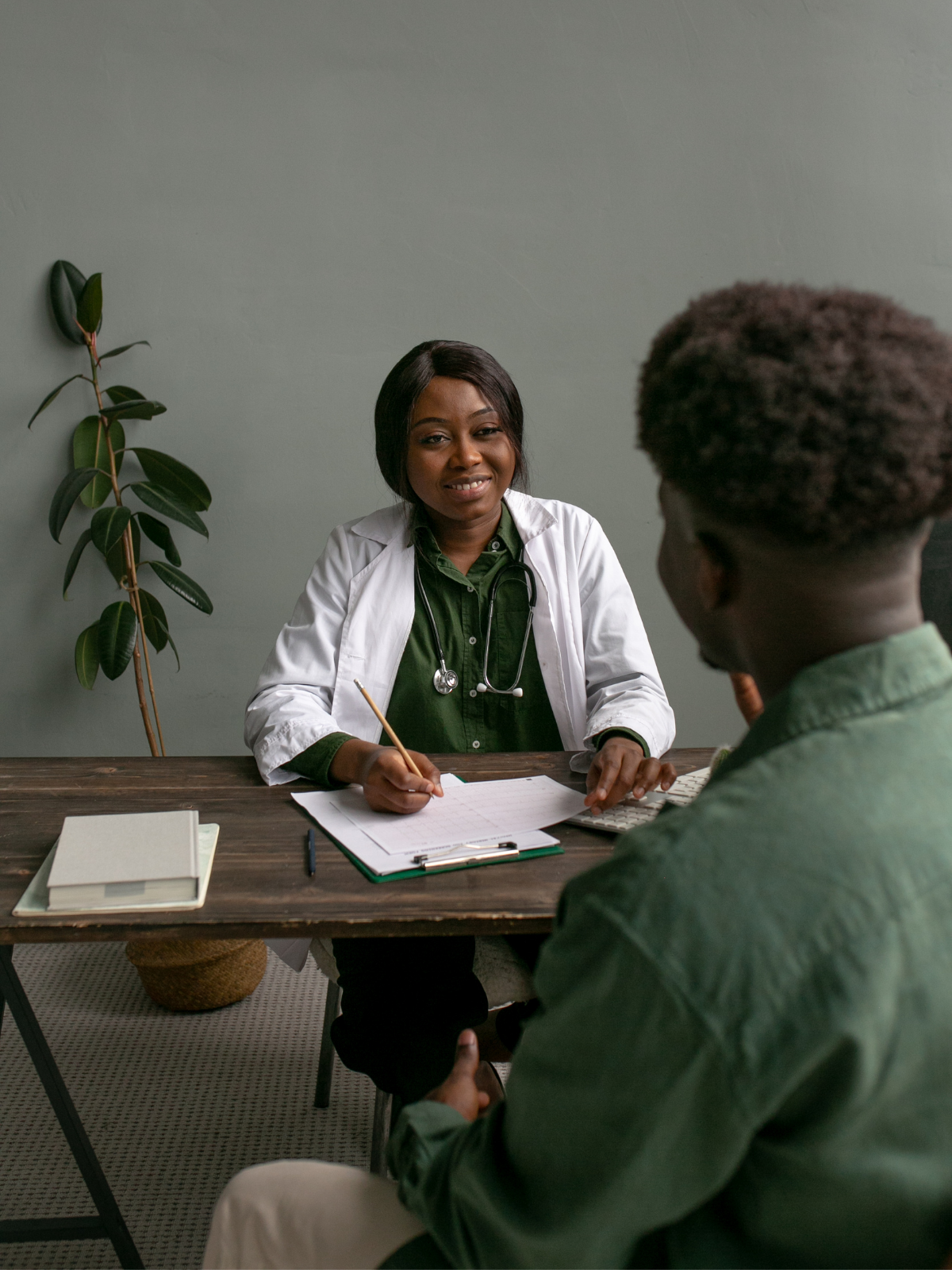 A female doctor with dark hair, wearing a white coat with a stethoscope, sitting at a wooden desk, talking to a male patient with dark hair and a green shirt, in an office with a green wall and a plant in the background.