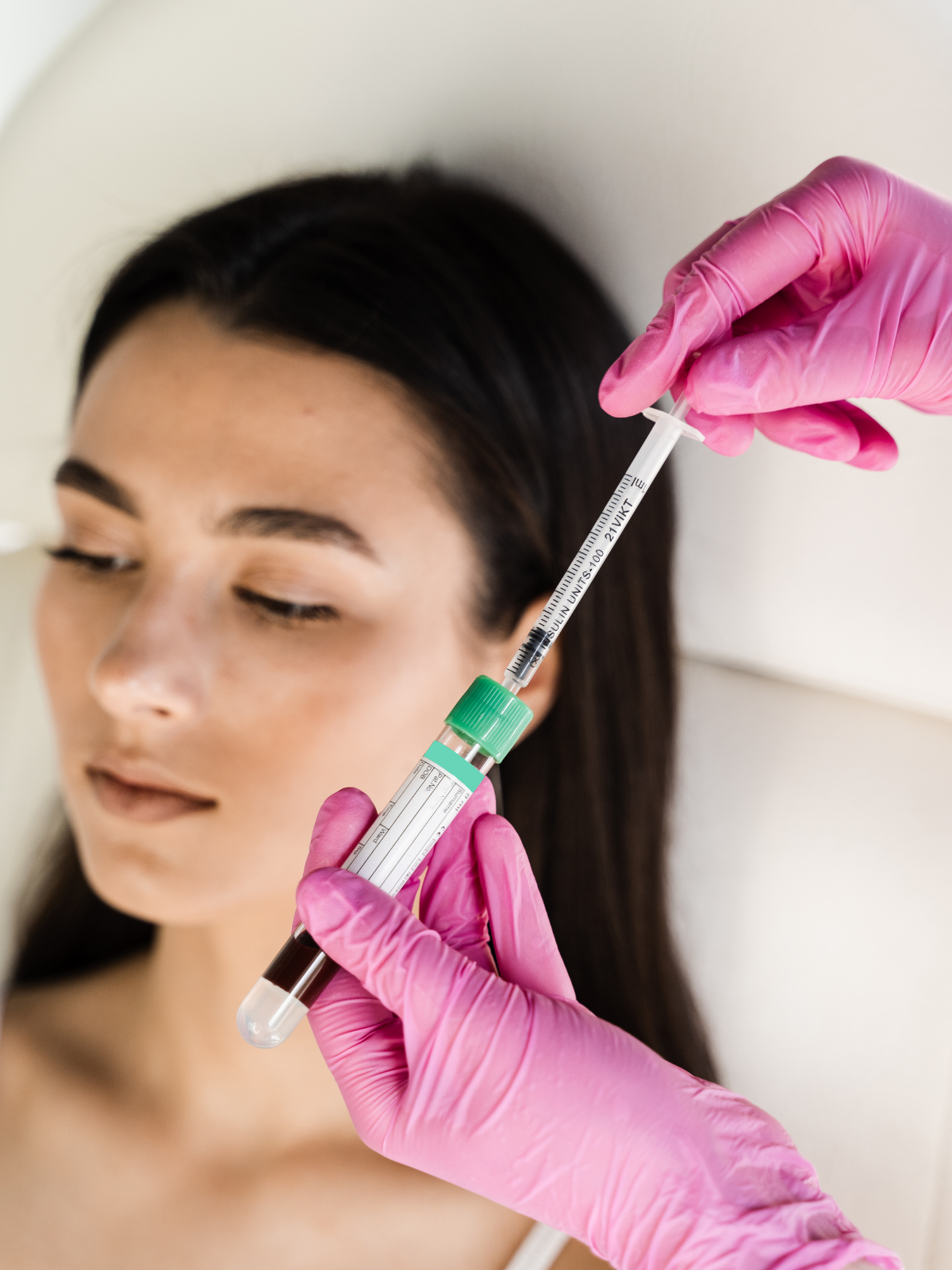 A woman with dark hair and closed eyes receives an injection in her face from a healthcare professional wearing pink gloves.