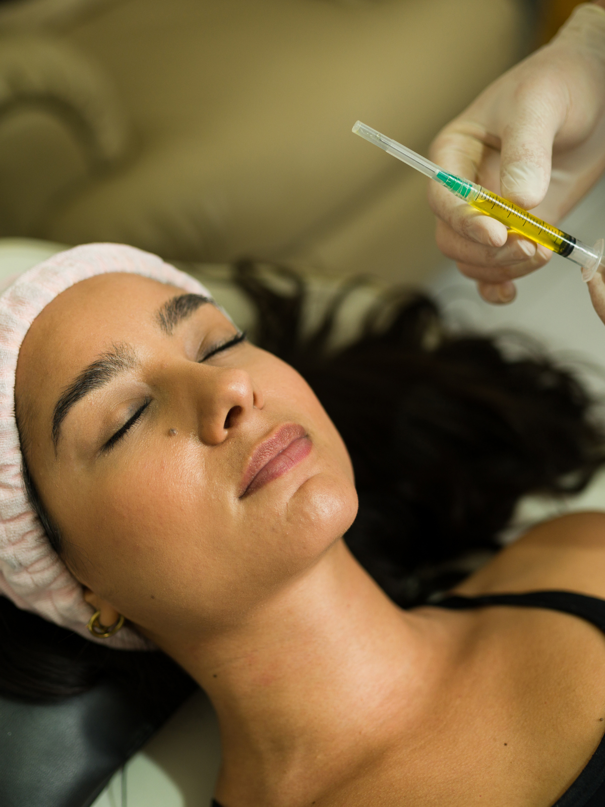 A woman is lying down with her eyes closed, receiving an injection from a healthcare professional in a clinical setting.