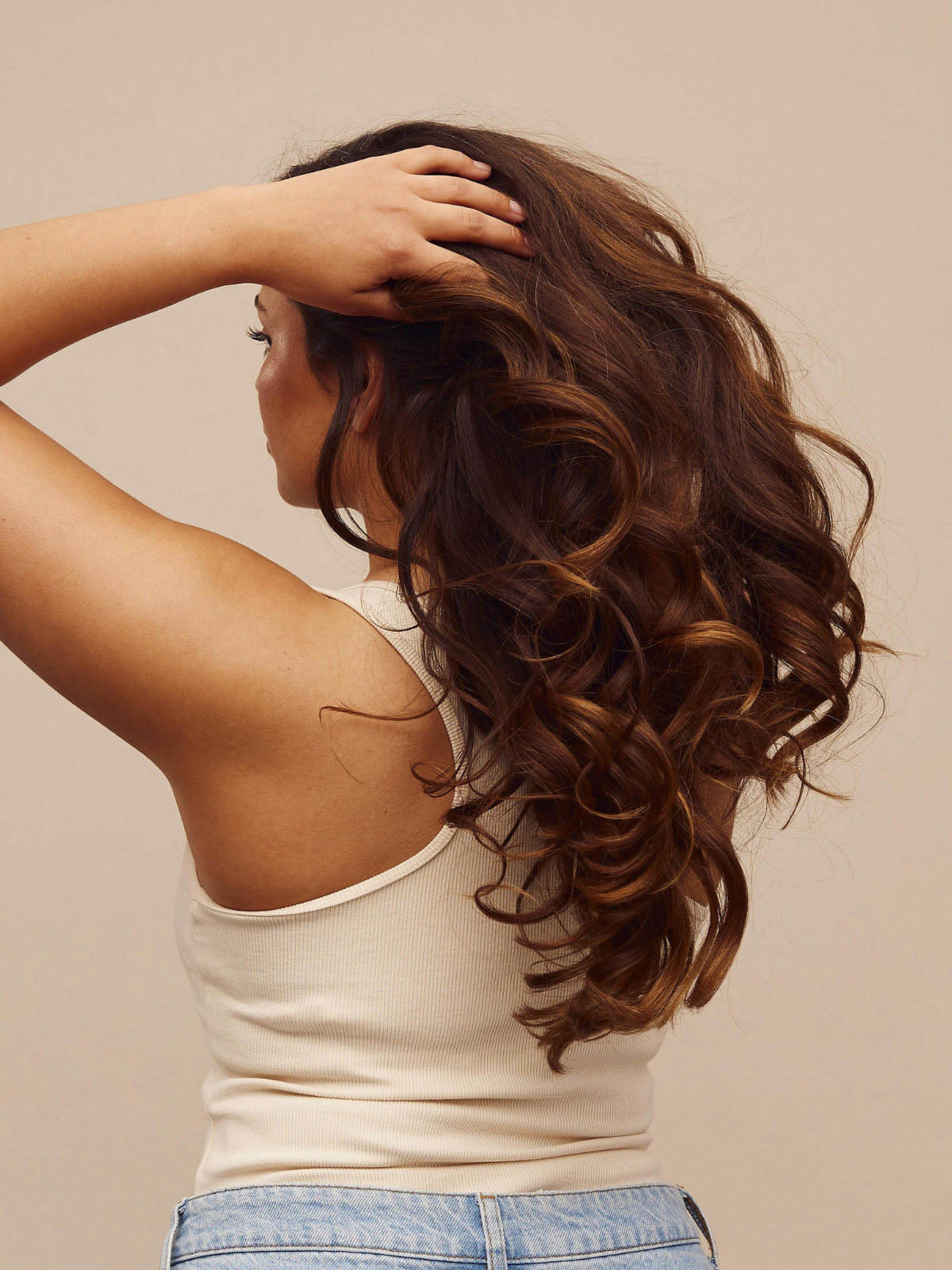 A woman with long, curly auburn hair wearing a beige tank top and denim shorts, posing with her hand on her head against a plain beige background.
