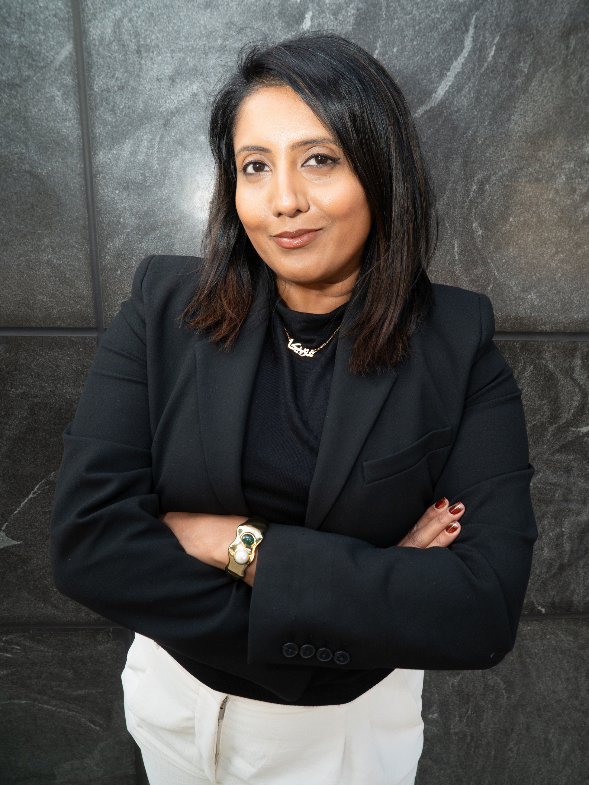 A woman with dark hair styled in a medium bob, wearing a black blazer, gold jewelry, and white pants, standing with crossed arms in front of a dark textured wall.