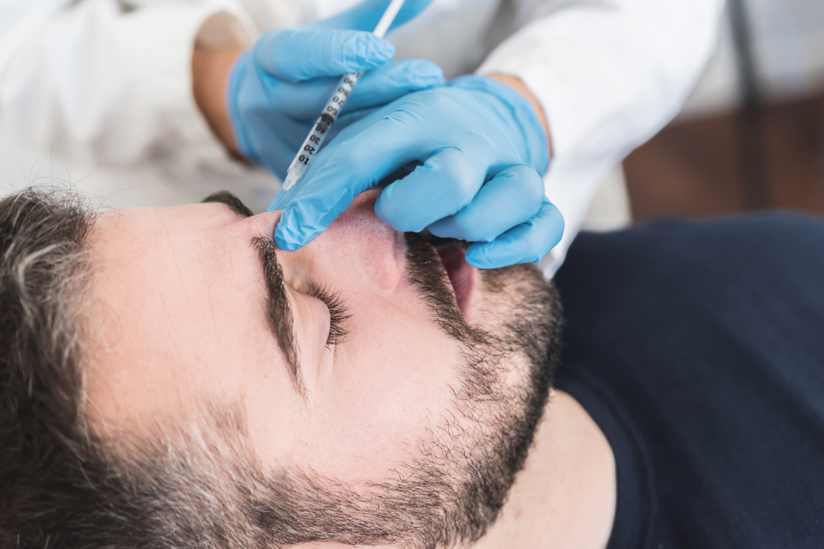 A healthcare professional in blue gloves administering a nasal injection to a man with a beard lying down.