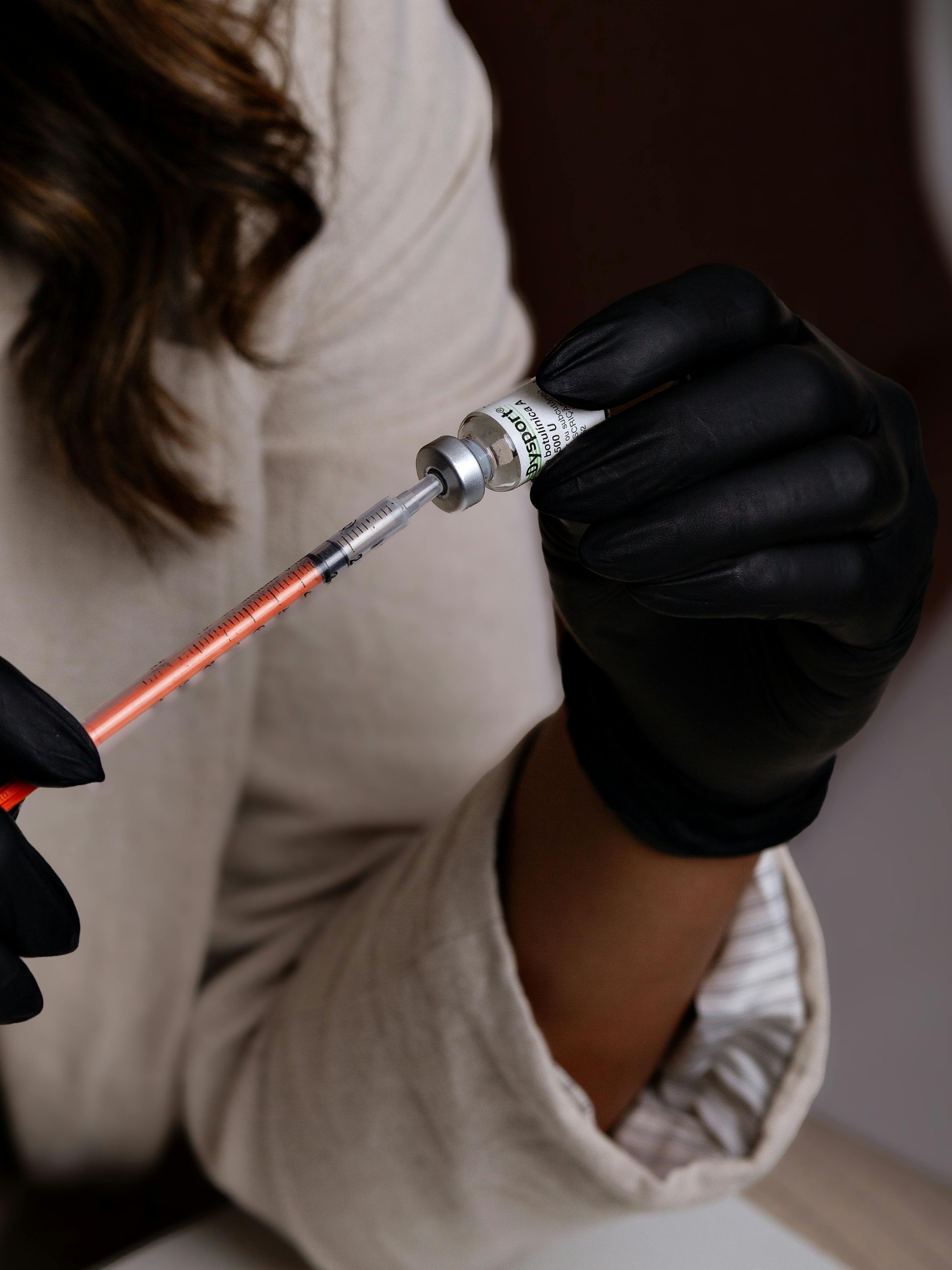 Person wearing black gloves drawing liquid from a vial with a syringe.