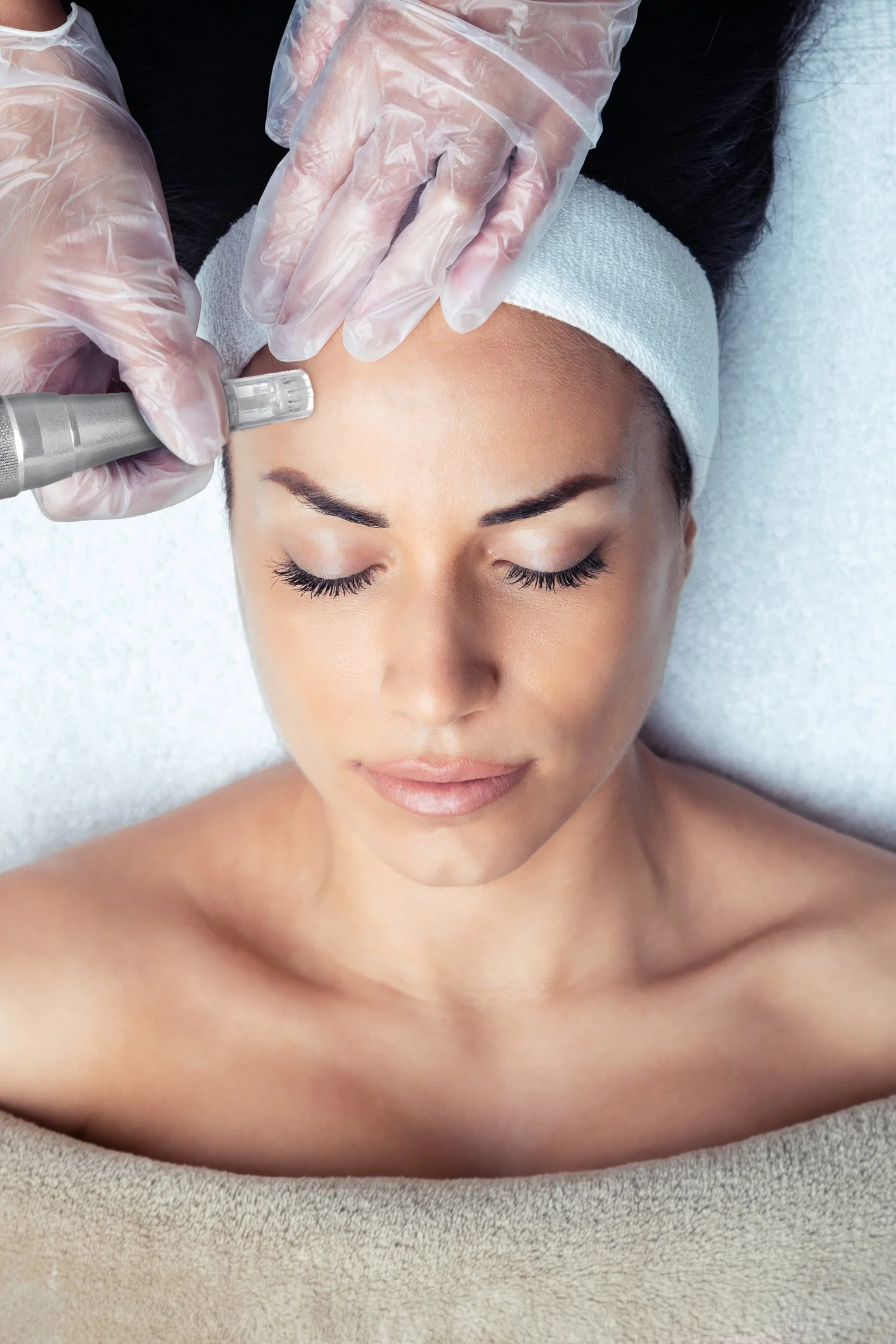 A woman is lying on a treatment table with her eyes closed, receiving a facial treatment. A practitioner wearing gloves is using a small tool on her forehead.