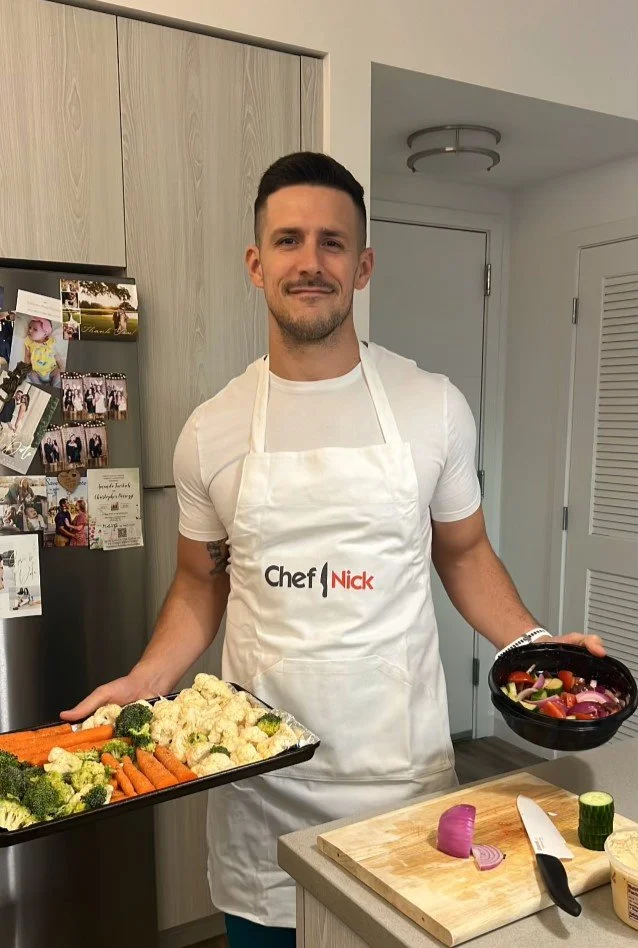 A man wearing a white apron with the logo 'Chef Nick' stands in a kitchen holding a tray of cooked vegetables including broccoli, cauliflower, and carrots, and a bowl of salad with sliced vegetables.