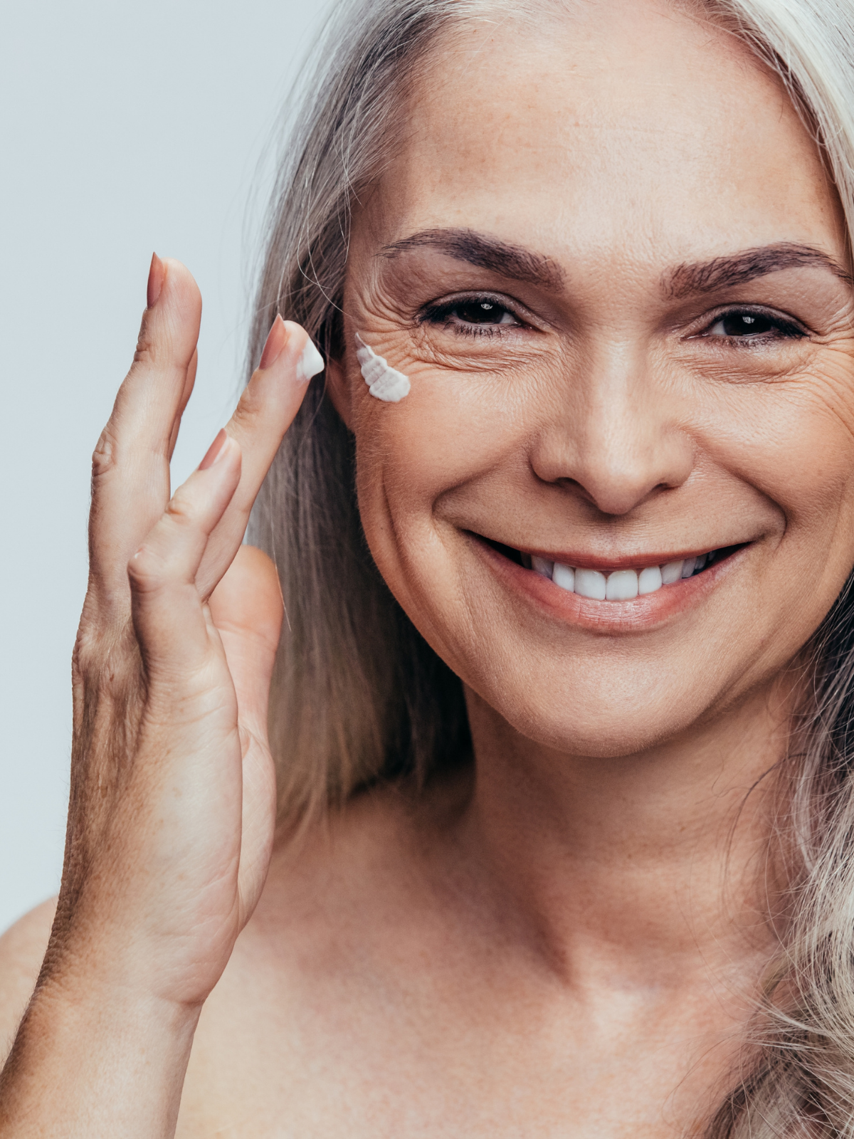 Close-up of a smiling mature woman with gray hair applying a skincare cream to her cheek.