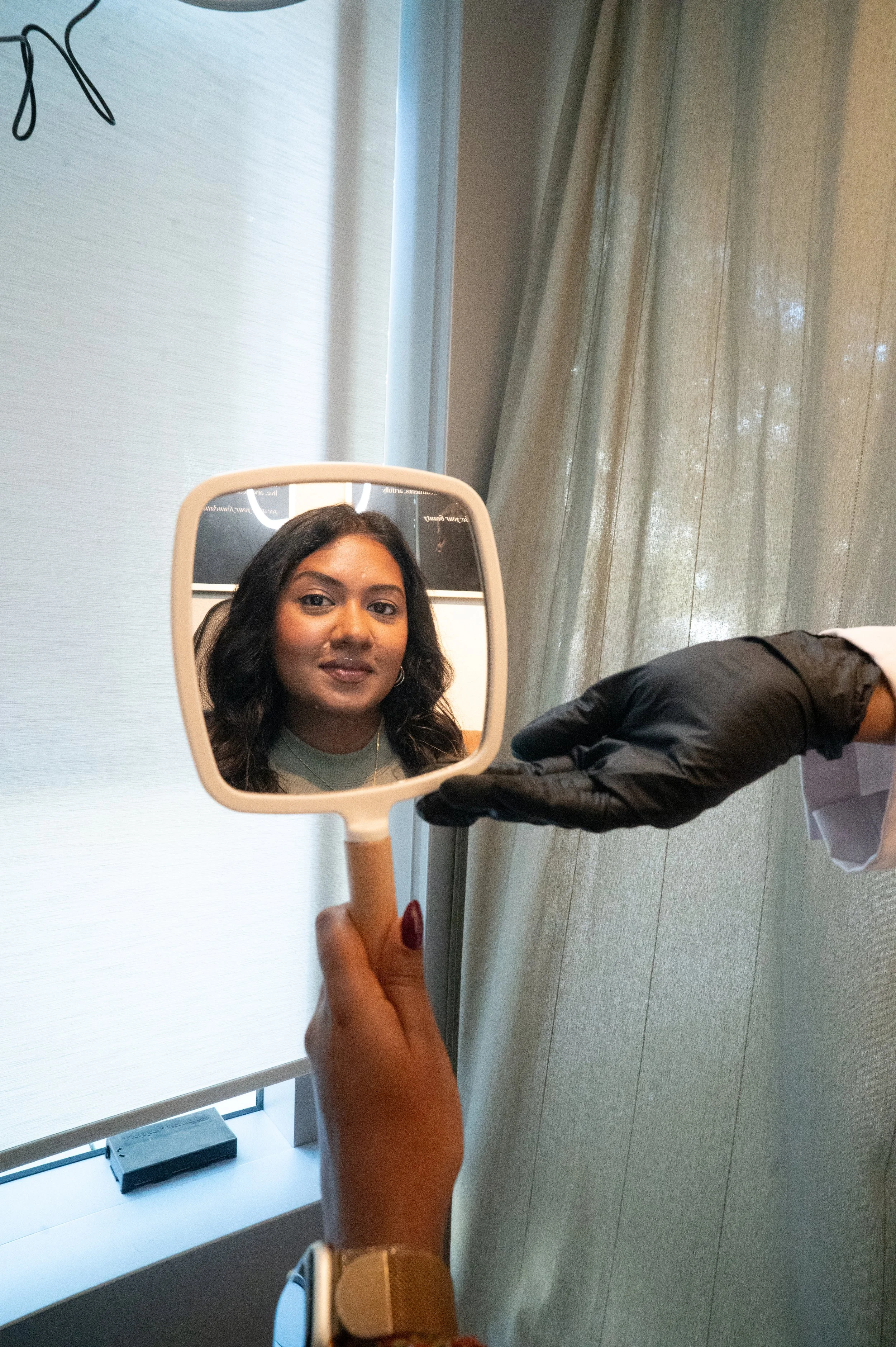 A woman is holding a small handheld mirror showing her face, with a person on the right side wearing a black glove. The scene appears to be indoors near a window with beige curtains.