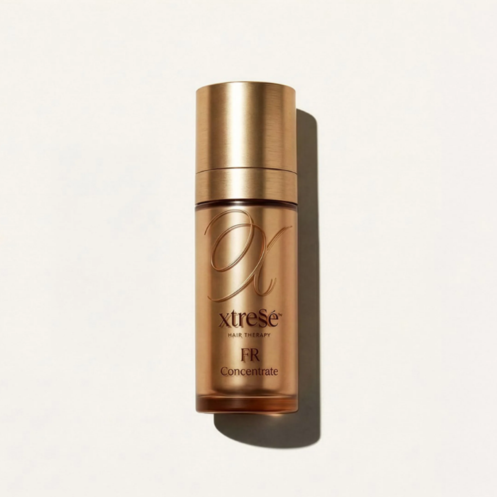 A rose gold bottle of Xtress hair therapy concentrate standing on a white surface, with a shadow cast behind it.