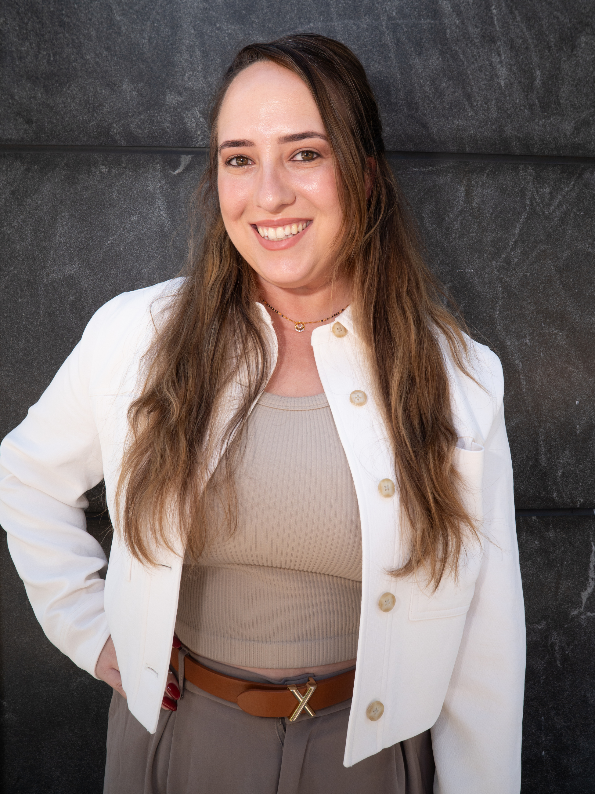 Portrait of a smiling woman with long brown hair wearing a beige top, white jacket, and brown belt, standing against a dark textured background.