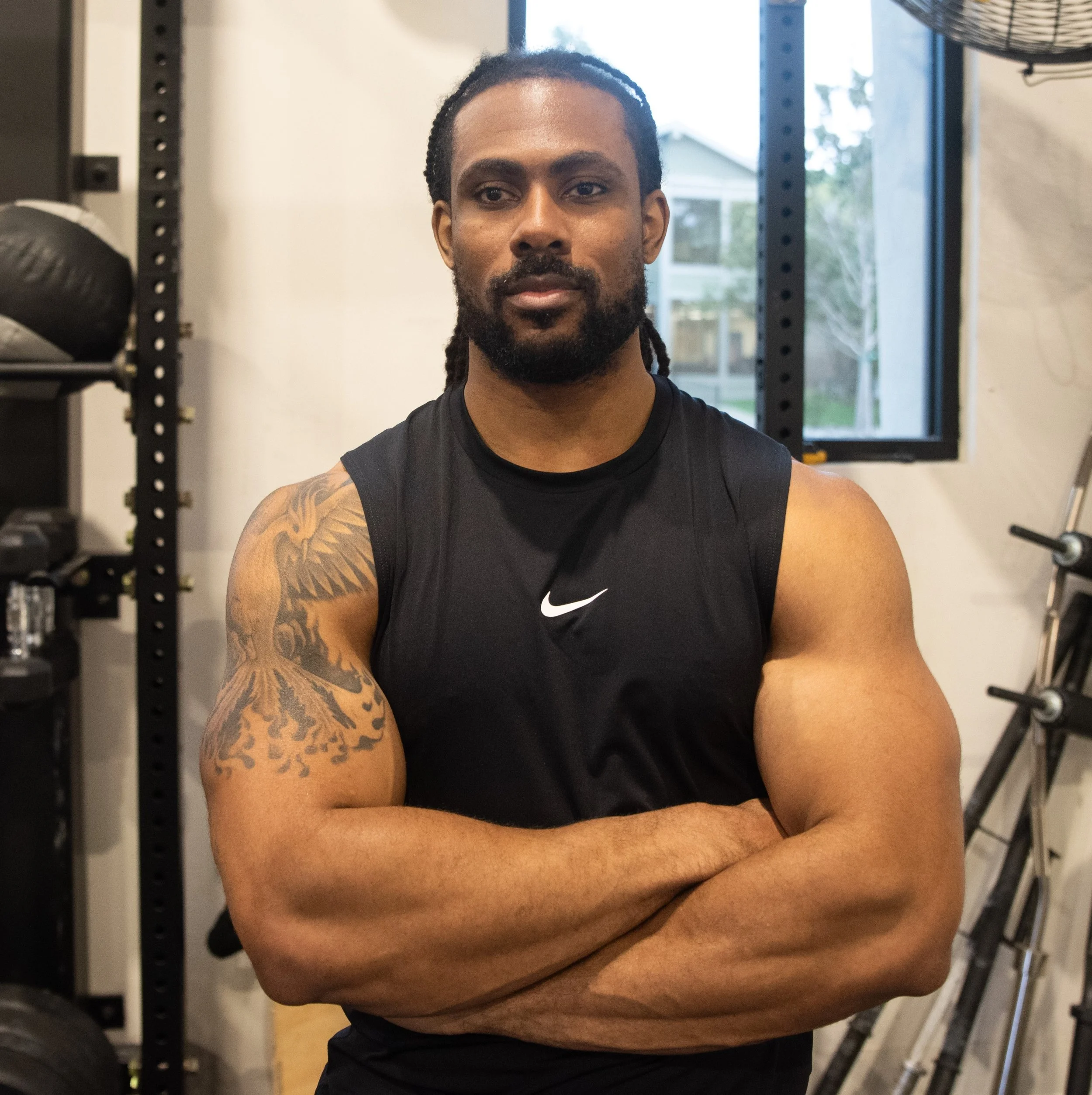 A man with dreadlocks and a beard taking a selfie in a gym, wearing a black Nike sleeveless shirt, with gym equipment and windows in the background.
