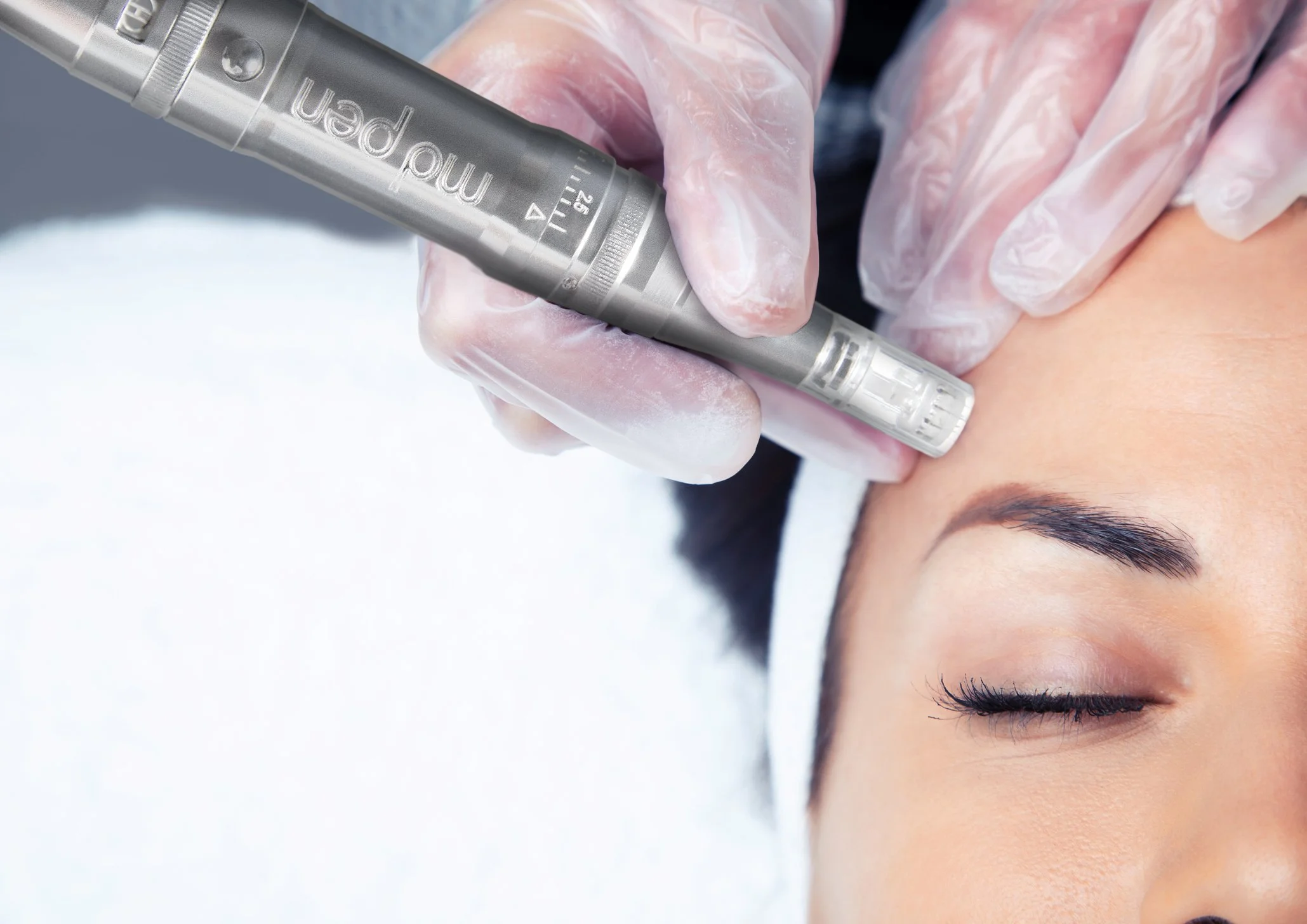 A close-up of a person receiving a cosmetic treatment or tattooing on their forehead with a metallic tool, while their eyes are closed.