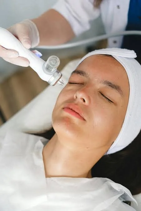 A person receiving a cosmetic or dermatological treatment on their face with a handheld device, lying down with eyes closed and wearing a white head covering.