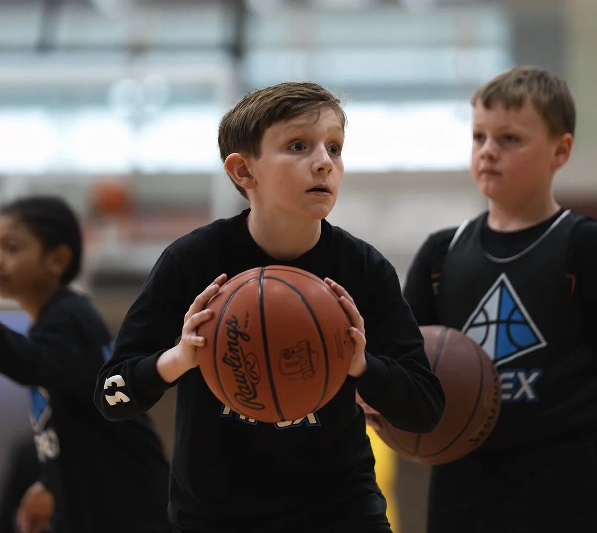 Young boy holding a basketball during a basketball practice or game, with other children in the background in an indoor gym.