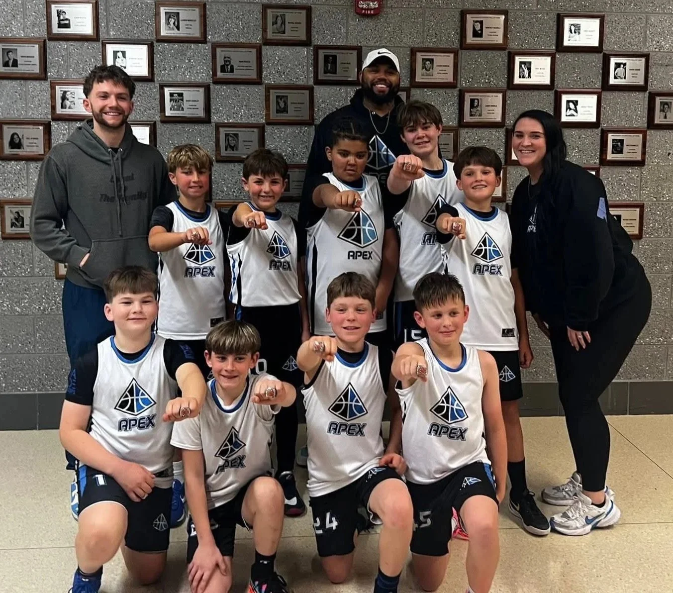 A group of young boys in basketball uniforms, along with two adults, posing together indoors against a wall with plaques. The boys wear white jerseys with a blue and black logo and the word 'APEX'. They are smiling and pointing at the camera with their fists.