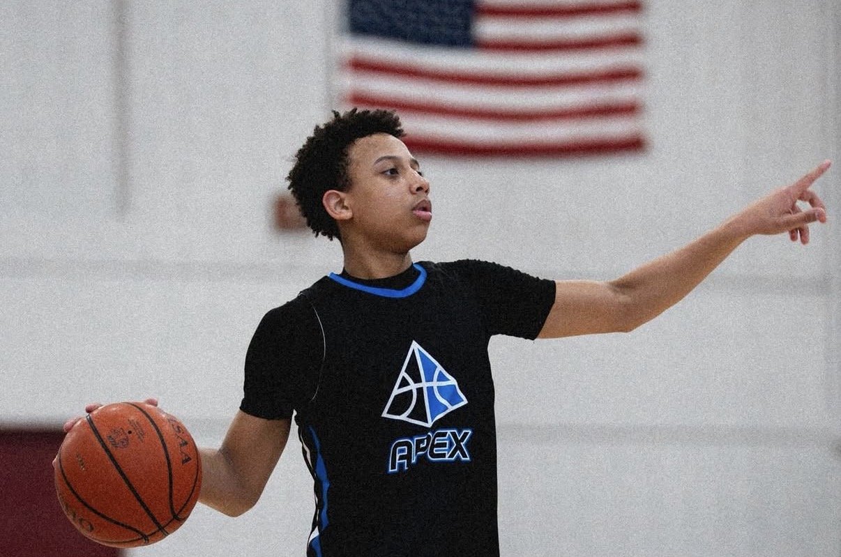 A young boy playing basketball indoors, wearing a black jersey with a blue and white logo that reads 'APEX', holding a basketball with his right hand, and pointing with his left hand, with an American flag in the background.