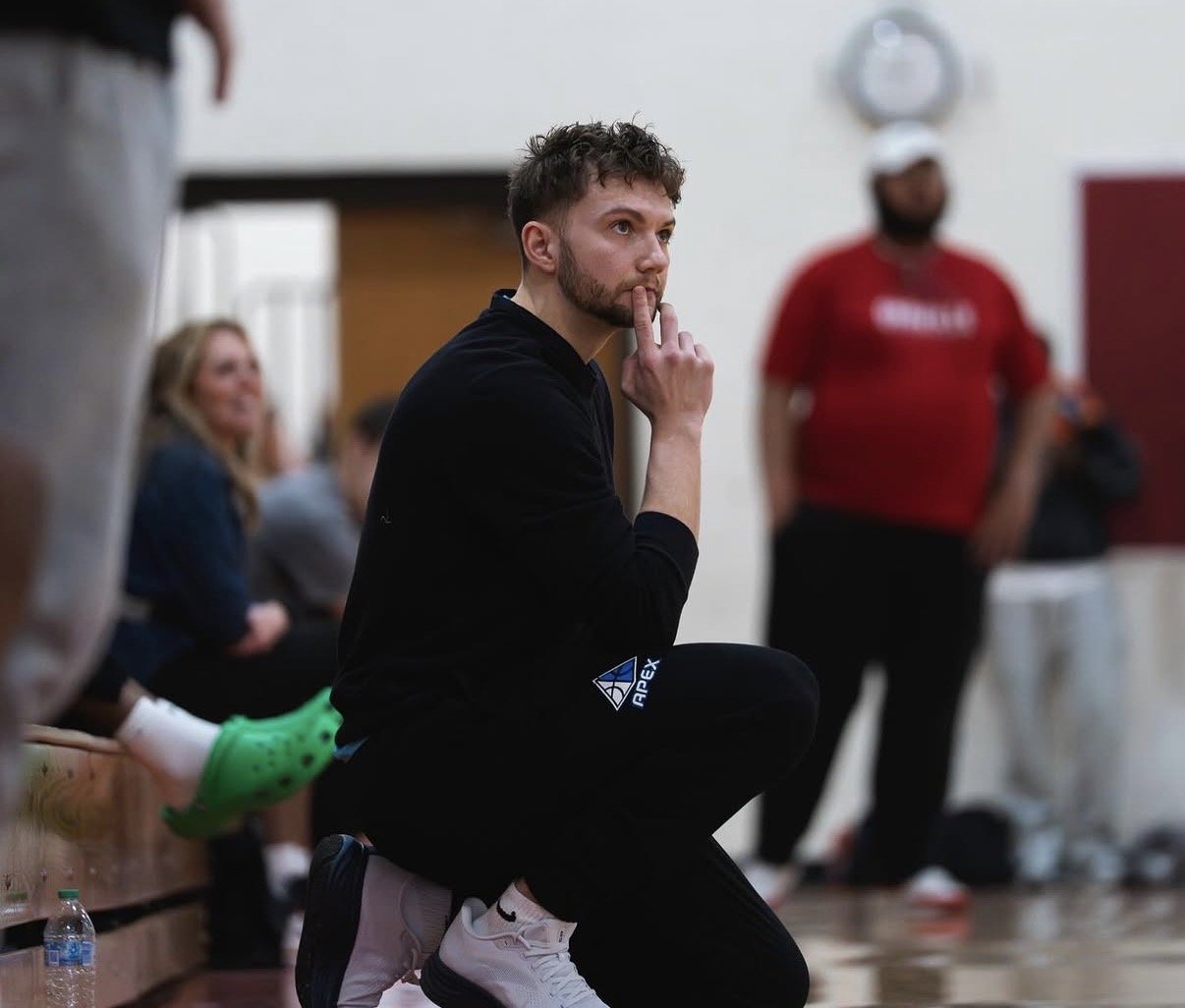 A young man with a beard and curly hair kneels on the floor, resting on his heels, with one finger on his lips. He is wearing a black long-sleeve shirt and black pants with a logo, and white sneakers. In the background, there are other people in a gym or indoor sports setting, including a woman sitting against the wall and a man wearing a red shirt and black hat, standing near a white wall with a clock.