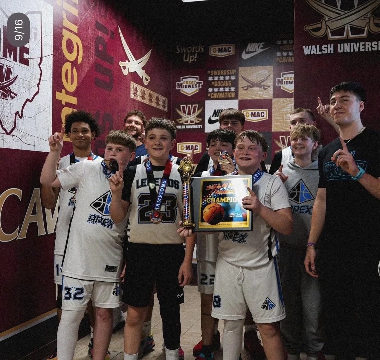 A group of young boys in athletic uniforms celebrating a basketball victory indoors, holding medals, a trophy, and a framed certificate that reads "Champions." They are smiling and pointing upward, with a backdrop featuring sports logos and banners.