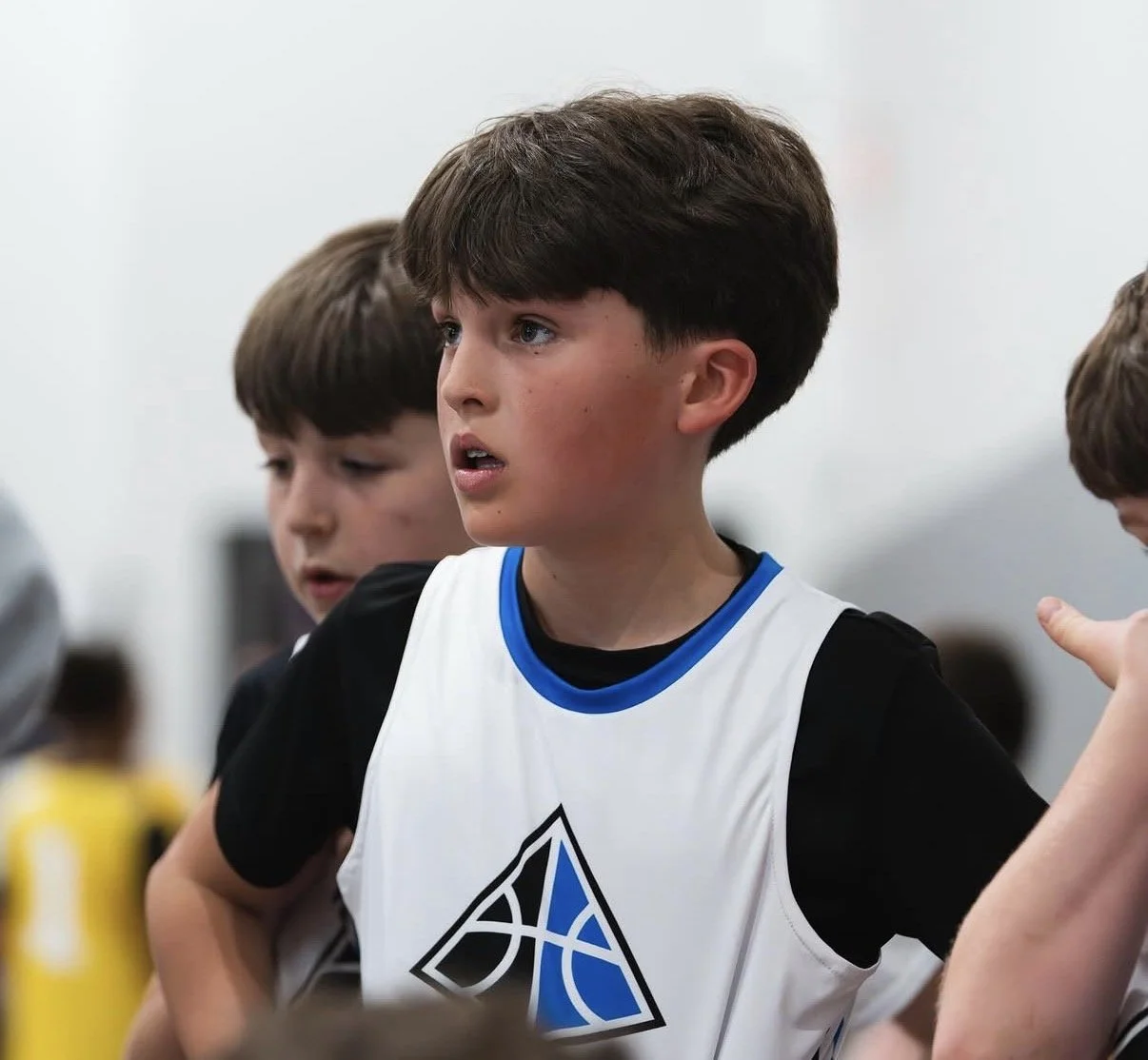 Young boy with brown hair and light skin wearing a basketball jersey with the Golden State Warriors logo, in a gym or sports facility, listening attentively.