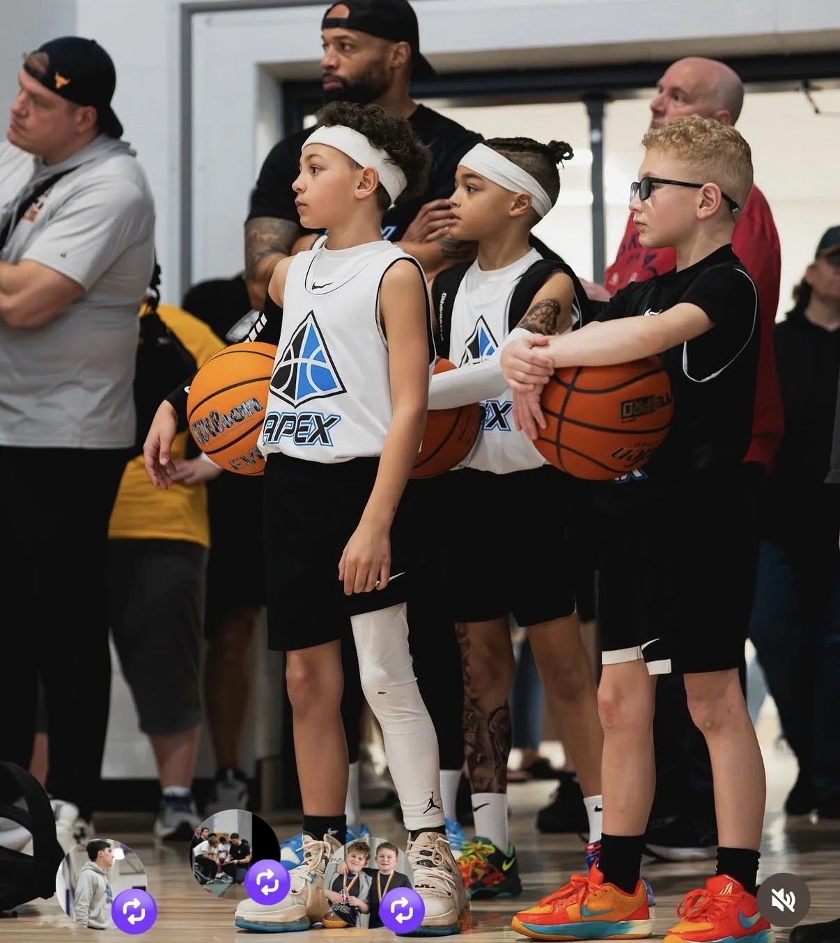 Young basketball players and adults standing in a gym, holding basketballs, with focused expressions. The children are wearing white jerseys with a logo and black shorts, while one has a white leg sleeve, and others have headbands and glasses. Adults are dressed casually, some with cross arms or hands over their hearts.