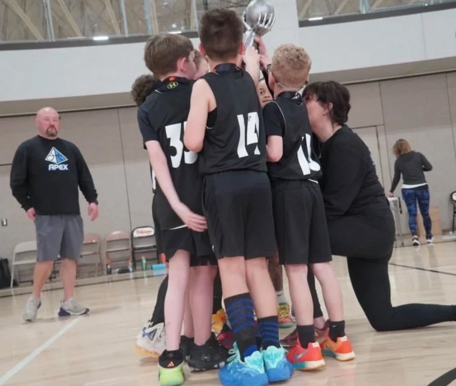 A youth basketball team in black jerseys with numbers is gathered in a huddle on an indoor court, holding a trophy while their coach kneels beside them. A man in a black hoodie stands nearby, and a woman and another person are in the background.