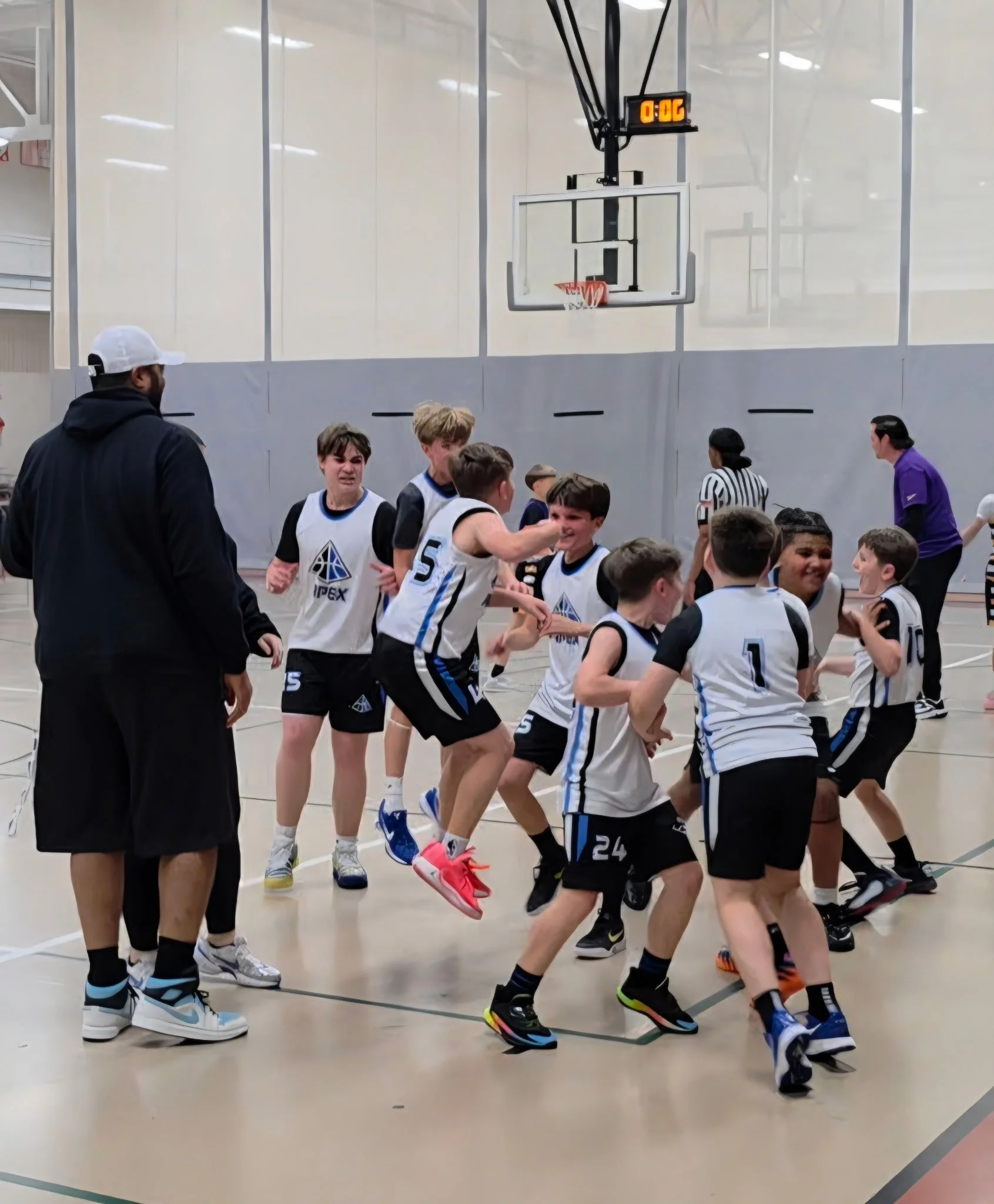 A group of young boys in basketball uniforms celebrating on an indoor basketball court. There is a basketball hoop and a digital scoreboard overhead. An adult in athletic wear is walking nearby.