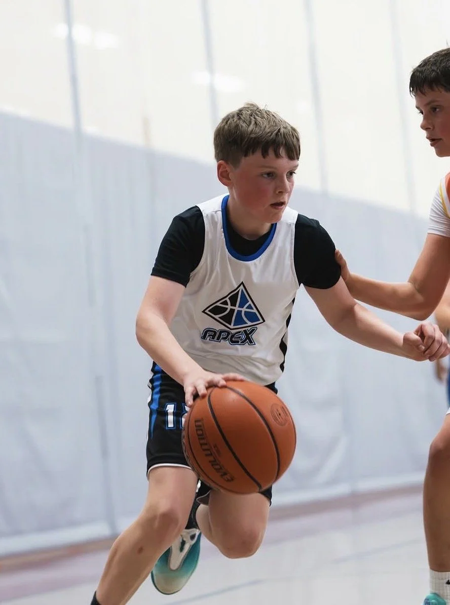 A young boy playing basketball on an indoor court, wearing a white and black uniform with an 'Apex' logo, dribbling a basketball as he is guarded by another player.
