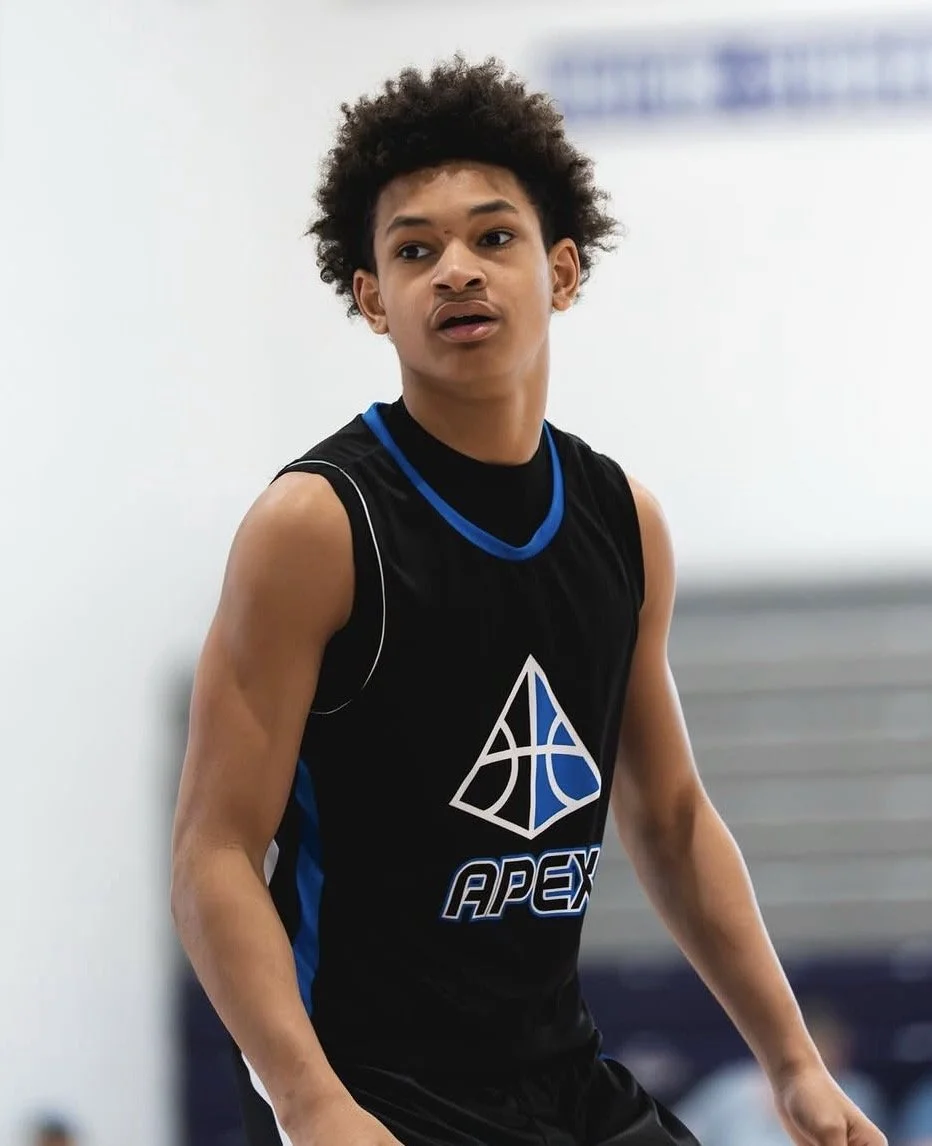 Young male basketball player in black sleeveless jersey with blue and white logo, standing indoors.