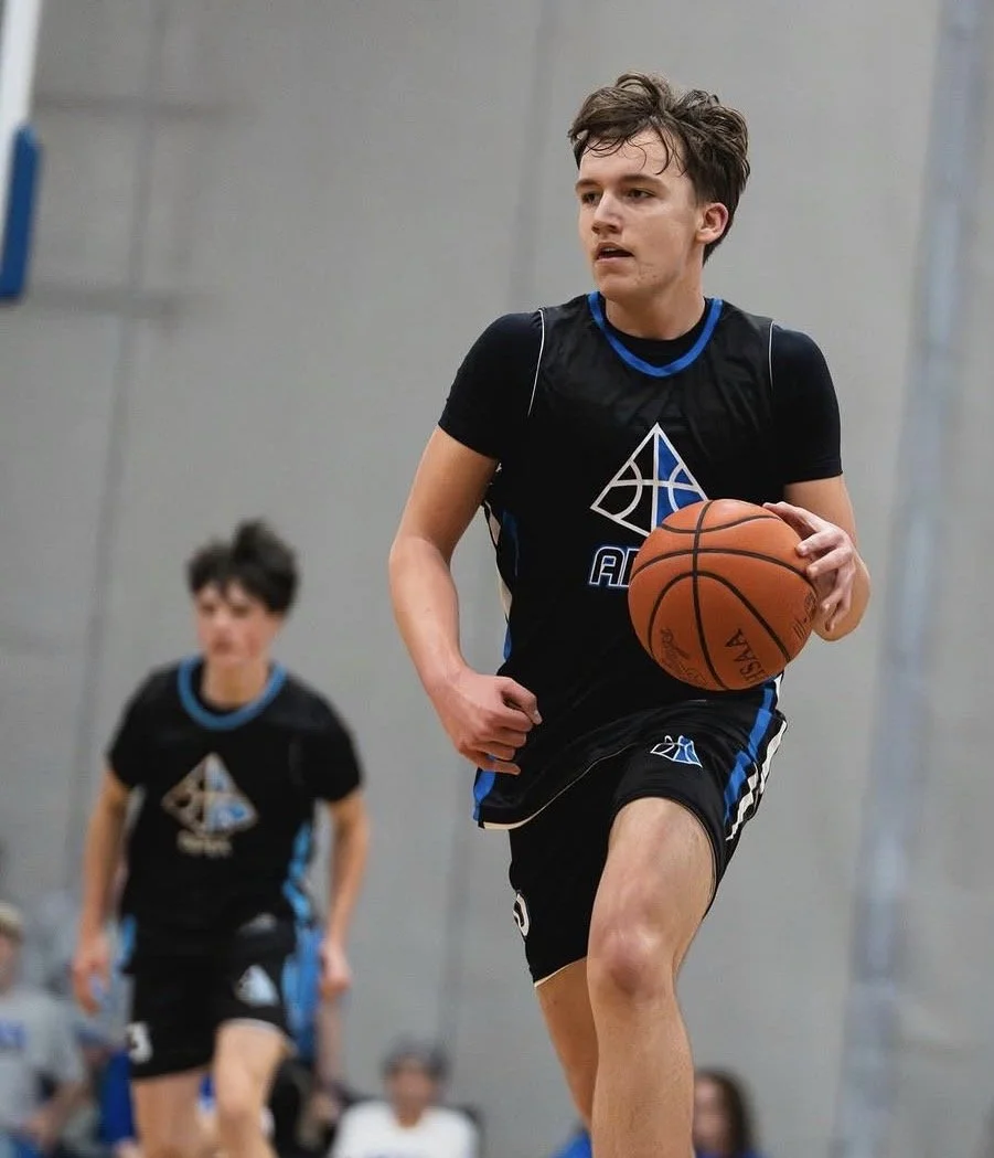 A young male basketball player dribbling a basketball on an indoor court with other players and spectators in the background.