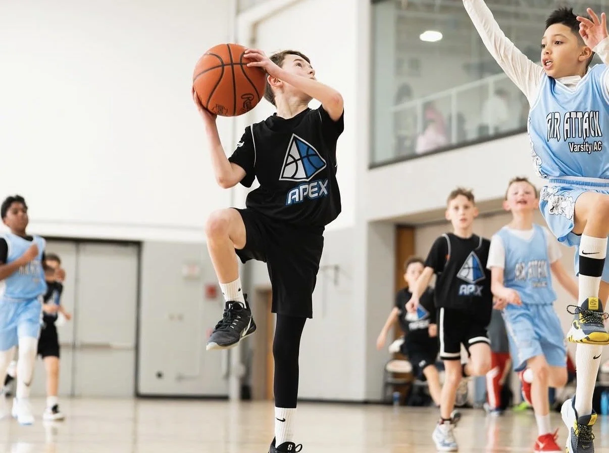 Young boys playing basketball in an indoor gym. One boy in a black jersey is jumping with a basketball near his face, while other children in jerseys watch or run in the background.