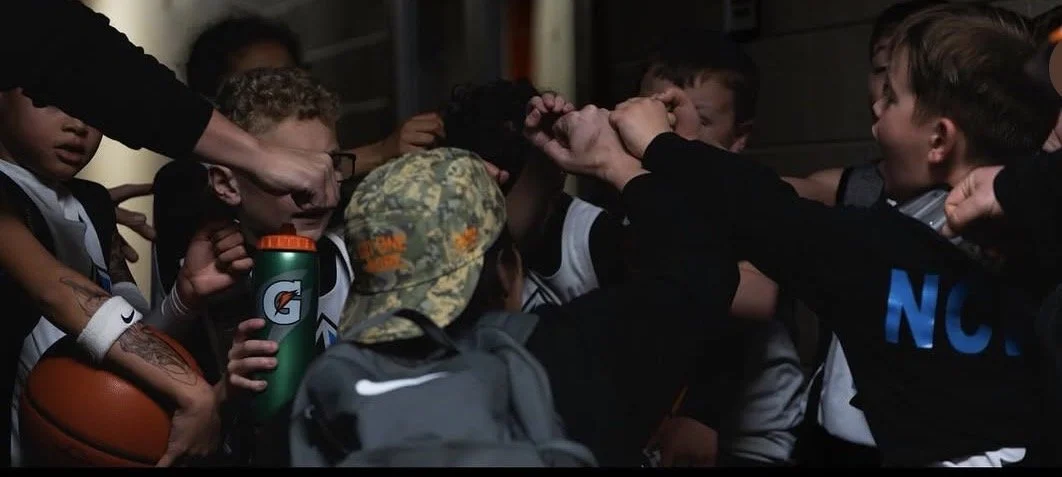 A group of young boys in a locker room huddle together with a coach, appearing to be in a team discussion or pep talk.