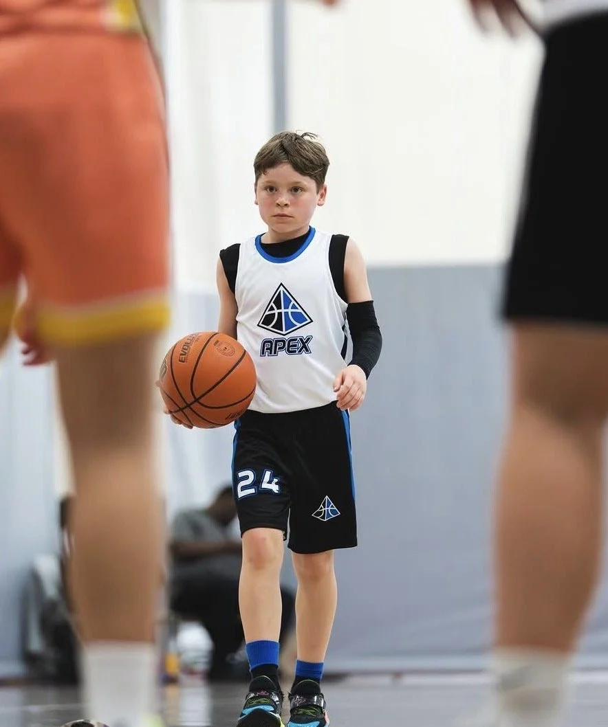 Young boy in a basketball uniform holding a basketball on an indoor court, surrounded by players.