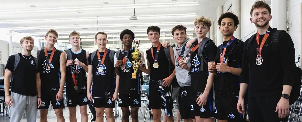 A group of ten young men in black sports uniforms and medals, standing together in a modern indoor space, holding a gold trophy and celebrating their victory.