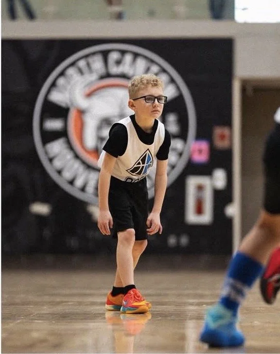 A young boy wearing glasses, a black and white sports jersey, black shorts, and bright orange sneakers, appears to be in a sports arena or gym, focused and ready to play basketball.