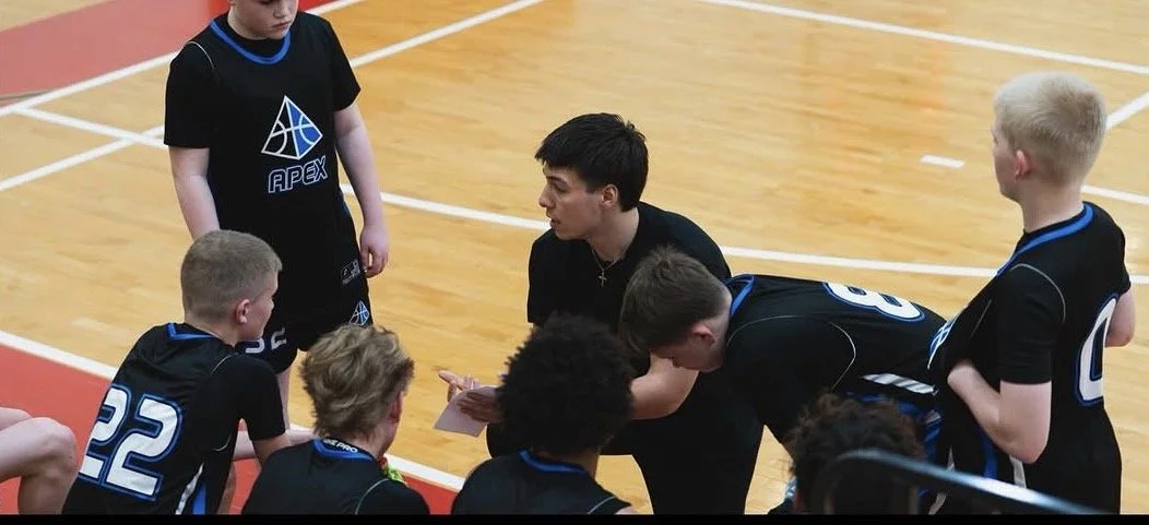 A sports team, wearing black jerseys with blue accents, gathered on an indoor basketball court listening to their coach, who is holding a piece of paper.