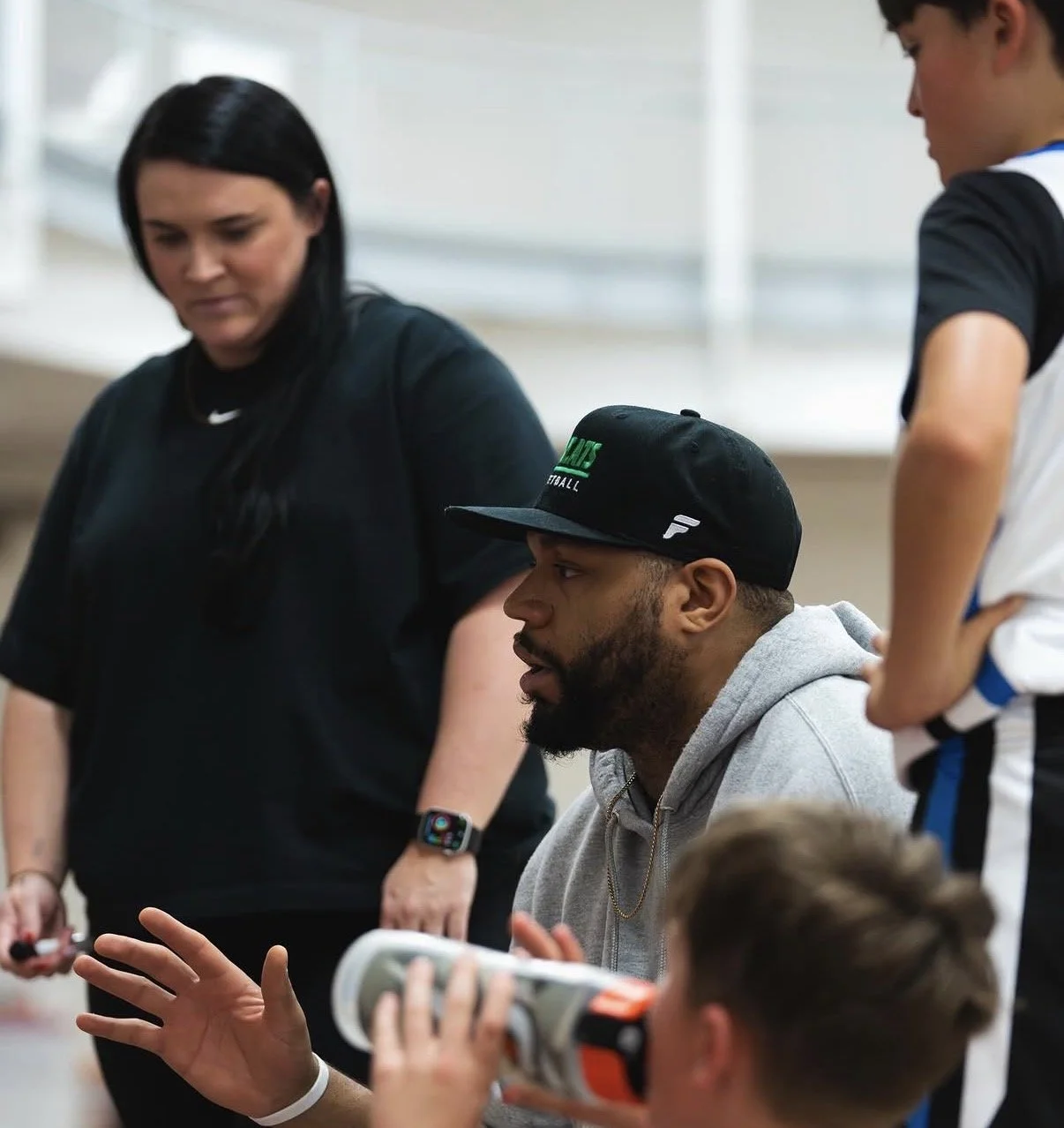 A man with a beard wearing a black cap and gray hoodie sitting and talking, as two children and a woman listen attentively in a bright room.