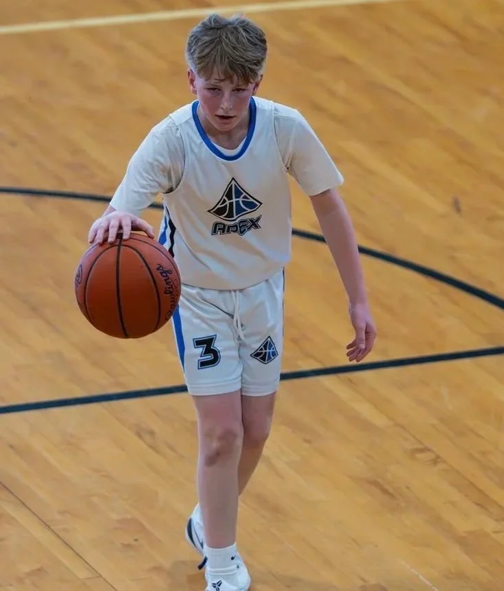 A young boy wearing a white basketball uniform with blue accents, holding a basketball, on a wooden basketball court.