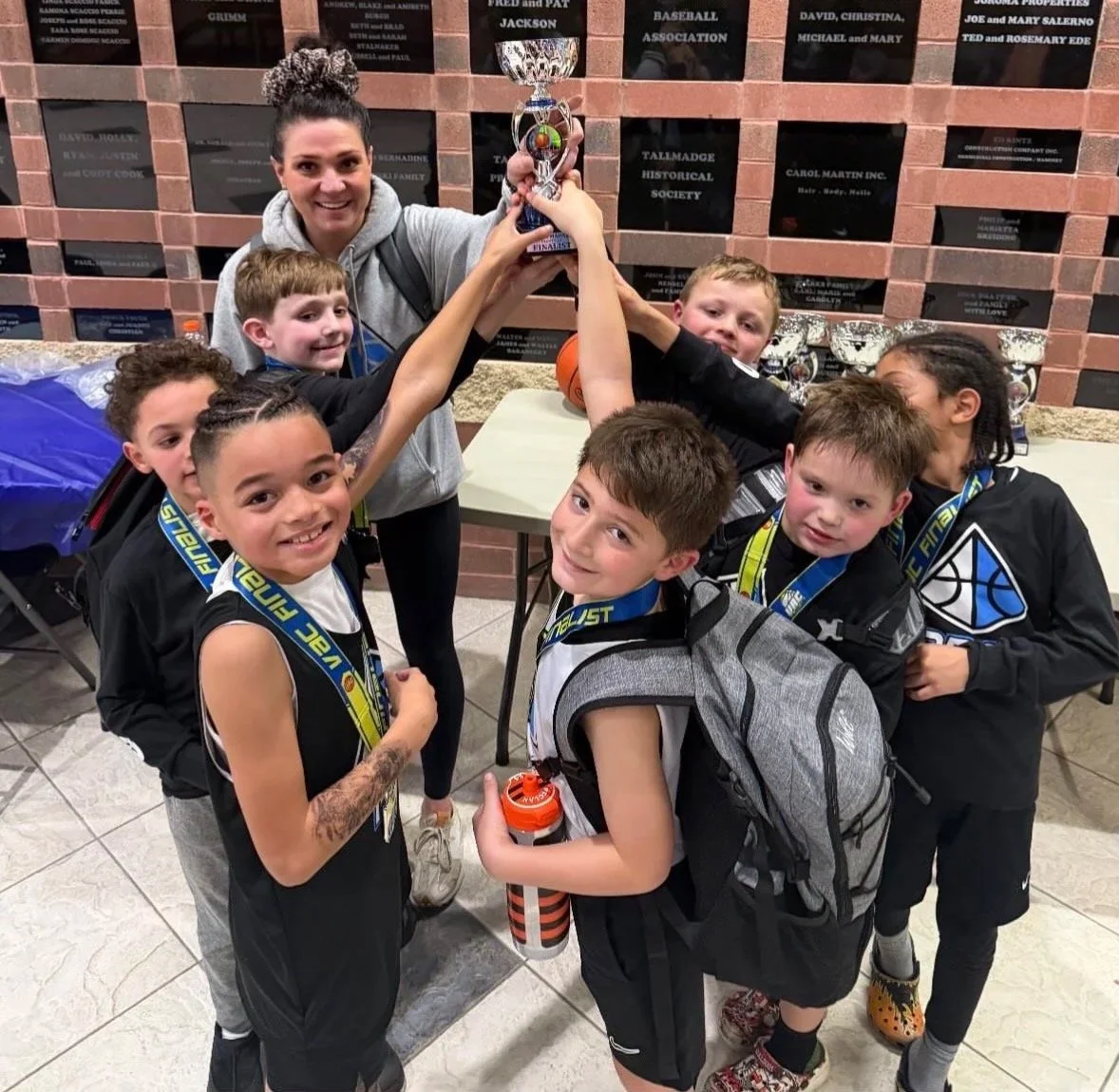 A group of children and a woman celebrating a victory, holding up a trophy together in an indoor setting with a wall of plaques in the background. The children are smiling, wearing sports attire and medals.