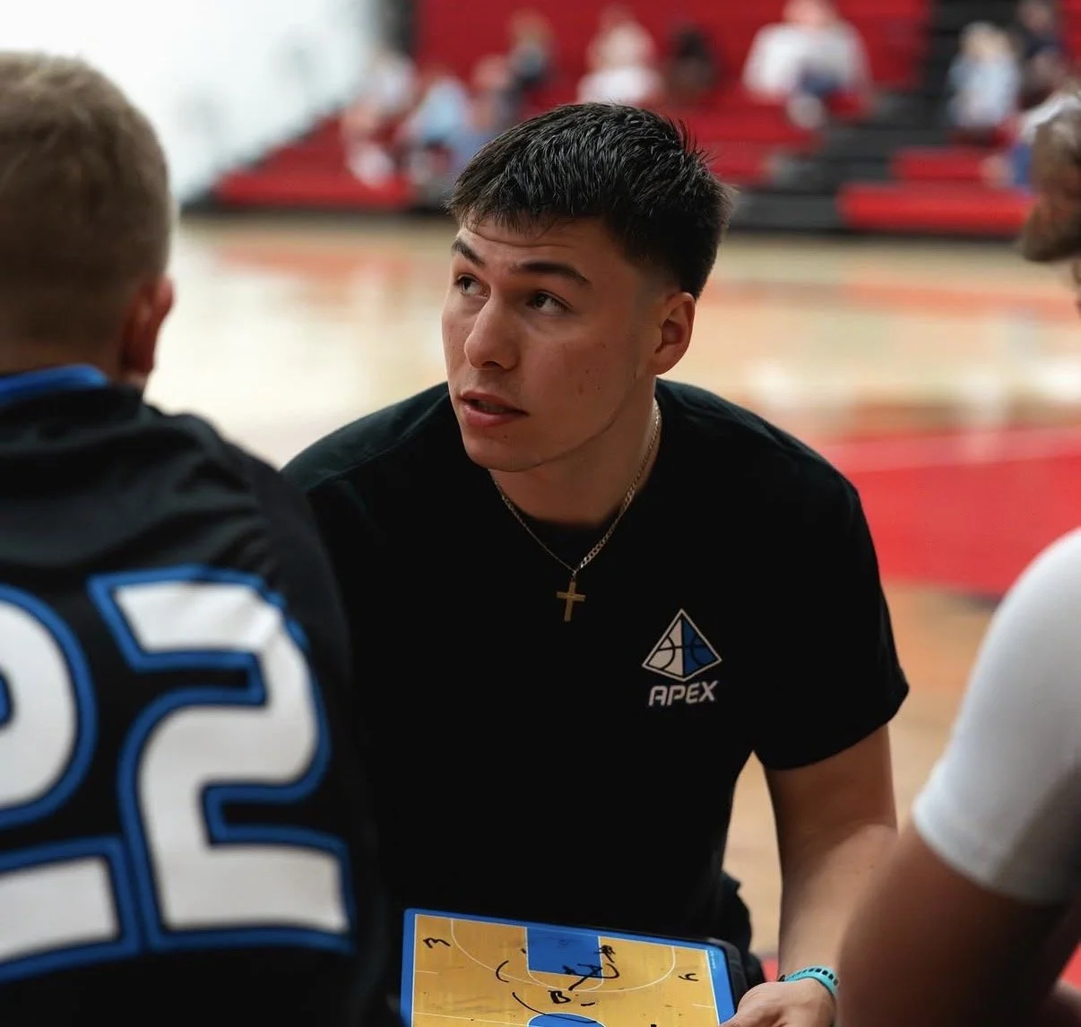 Young man coaching basketball at a game in a gymnasium with players and spectators in the background.
