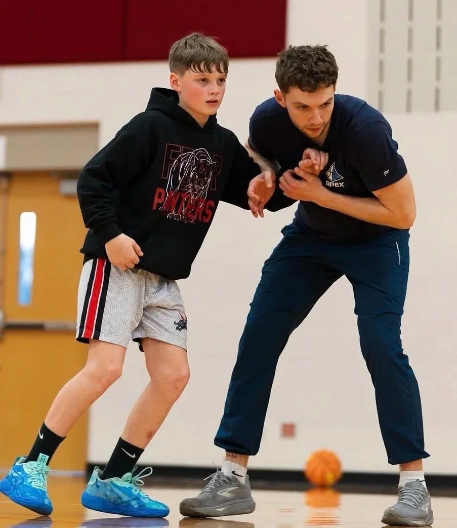 A young boy and a man are practicing wrestling on a gymnasium floor. The boy is wearing a black hoodie, gray shorts, and bright blue sneakers. The man is dressed in a navy polo shirt and navy pants, demonstrating a wrestling hold with the boy. There is a basketball in the background.
