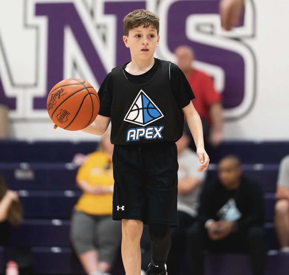 Young boy playing basketball indoors, wearing a black jersey with a blue and white logo and the word "APEX" on it, holding a basketball, with a purple and white wall and spectators in the background.