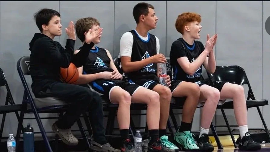 Four young boys in basketball uniforms sitting on a bench in a gym, reacting to the game; some are clapping and cheering.