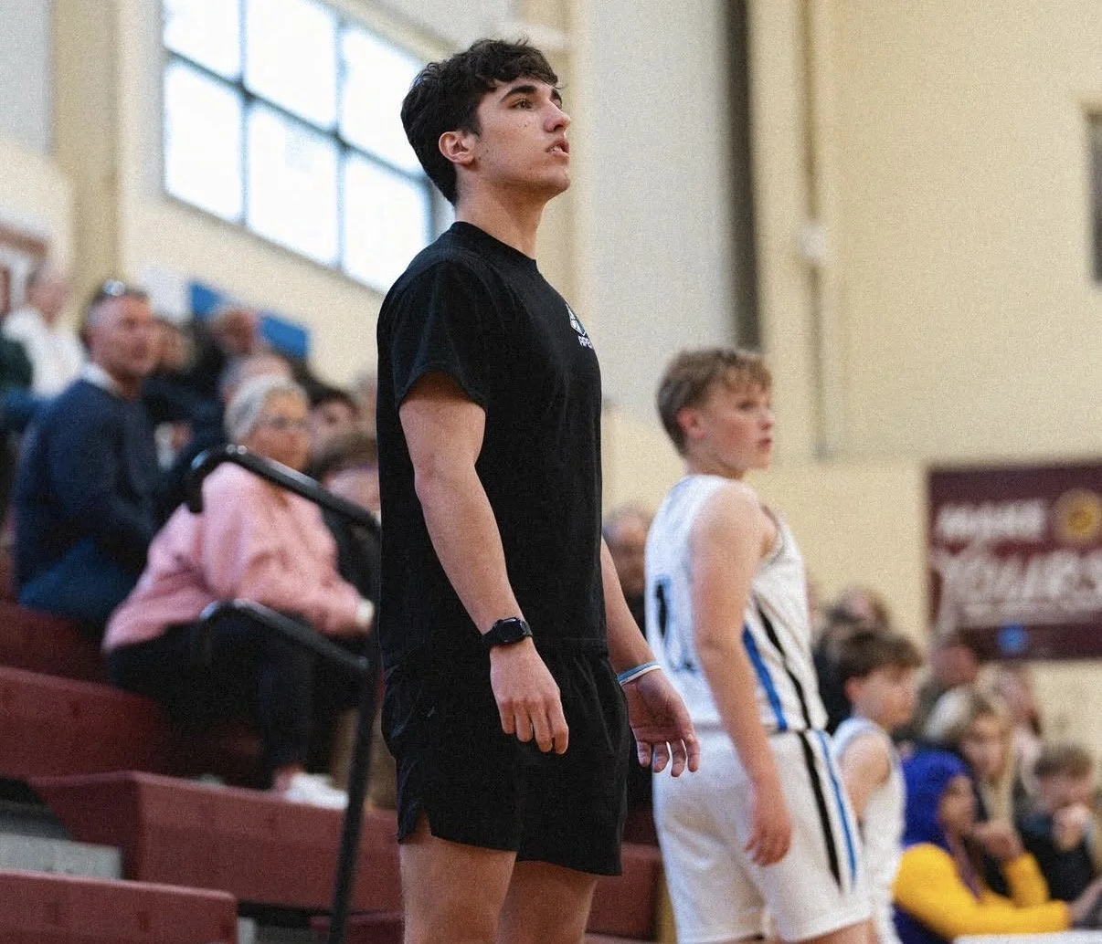 Young male basketball player standing on the court during a game, wearing a black uniform, with spectators watching from the bleachers behind him.